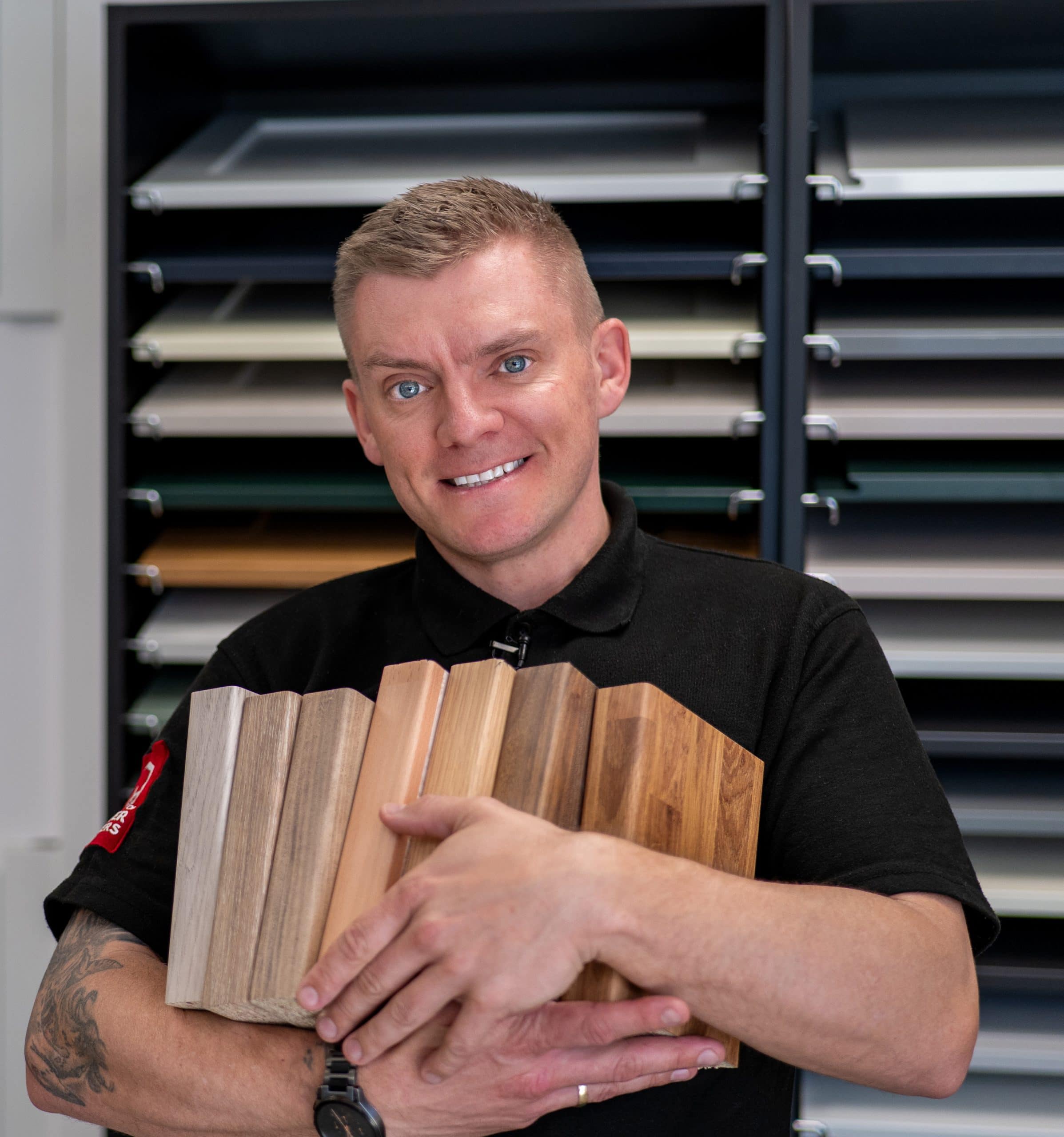A smiling man in a black shirt holds several wood samples in his arms, standing in front of shelves displaying various coloured panels in a kitchen and bathroom showroom.