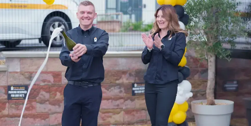 A smiling man in a black shirt pops a bottle of champagne as foam sprays out, while a woman in a black outfit claps. They celebrate outdoors near a stone wall and black and yellow balloons after a successful kitchen and bathroom showroom installation.