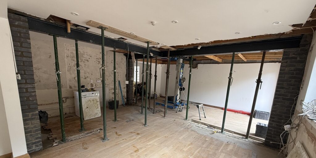 A room under refurbishment with exposed support beams and metal poles holding up the ceiling; brick walls and unfinished surfaces are visible, reflecting a space in progress for kitchen and bathroom showroom installation. Tools and building materials are scattered around.