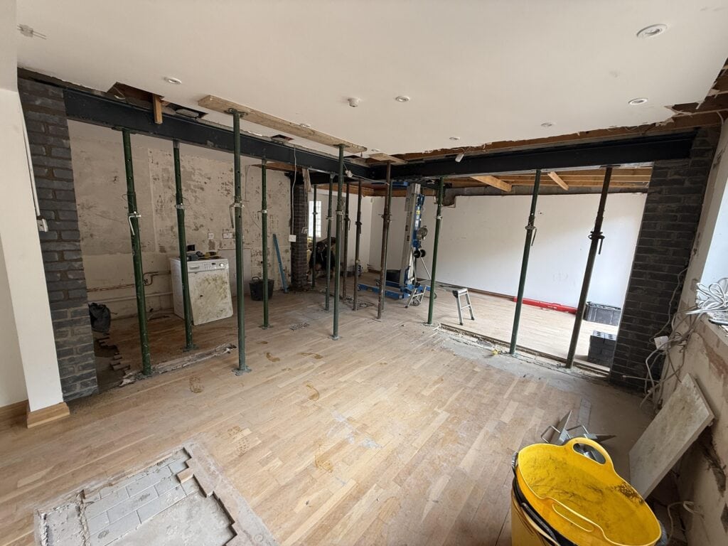 A room undergoing renovation with exposed brickwork, steel support beams, and metal poles propping up the ceiling—ideal for a future kitchen and bathroom showroom installation. The floor is partially removed, with tools, debris, and a yellow bucket scattered about.