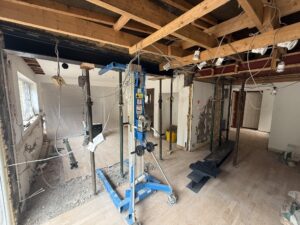 A house interior under renovation with exposed wooden beams, wiring, and support columns hints at future kitchen and bathroom showroom installation. Construction equipment and debris scatter the floor as natural light enters through a window on the left.