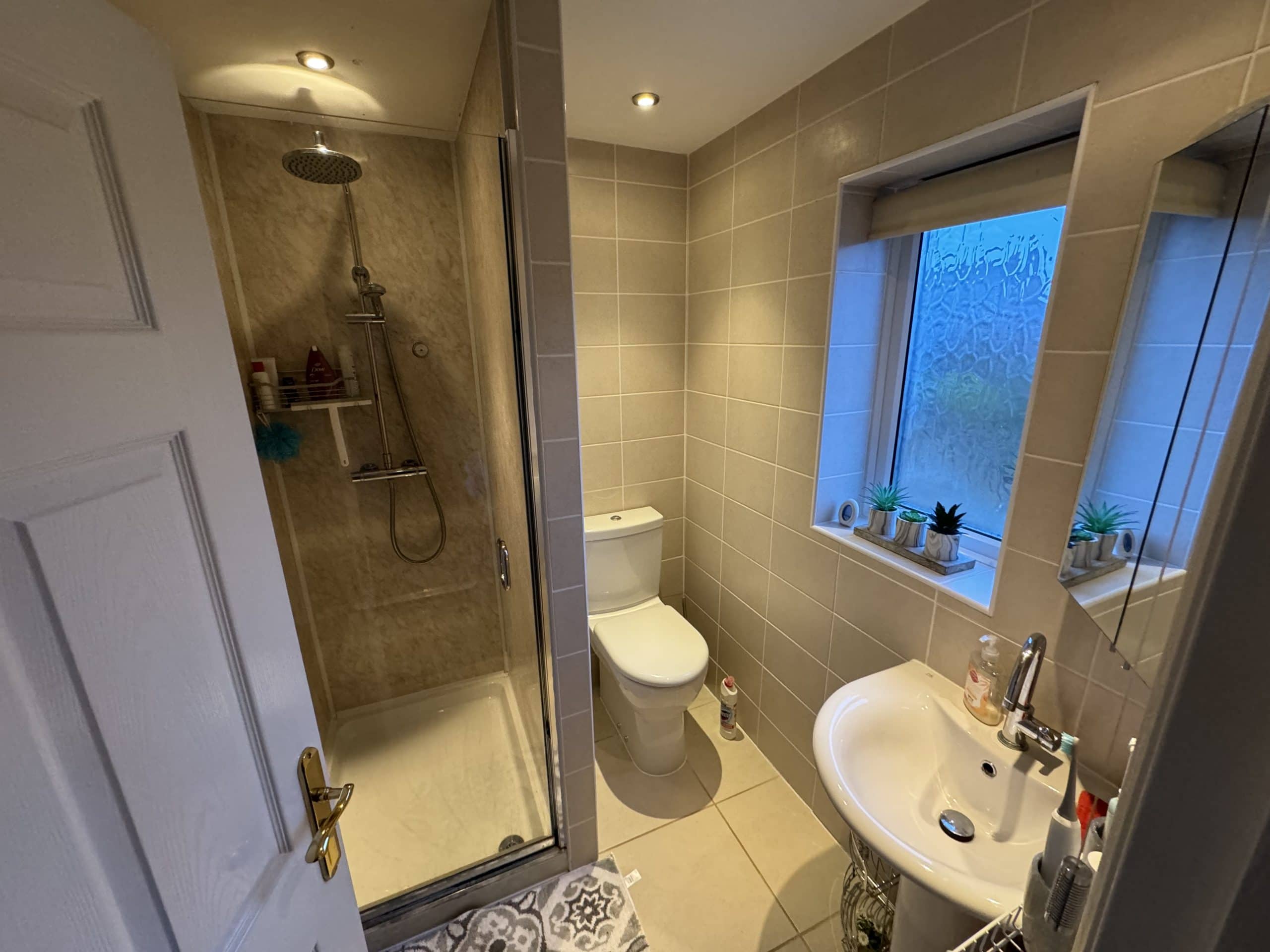 A modern bathroom designed with beige tiles, featuring a sleek glass shower on the left, a central toilet, and a white sink with mirror. Several potted plants decorate the windowsill and shelf. Ideal for kitchen and bathroom showroom inspiration.