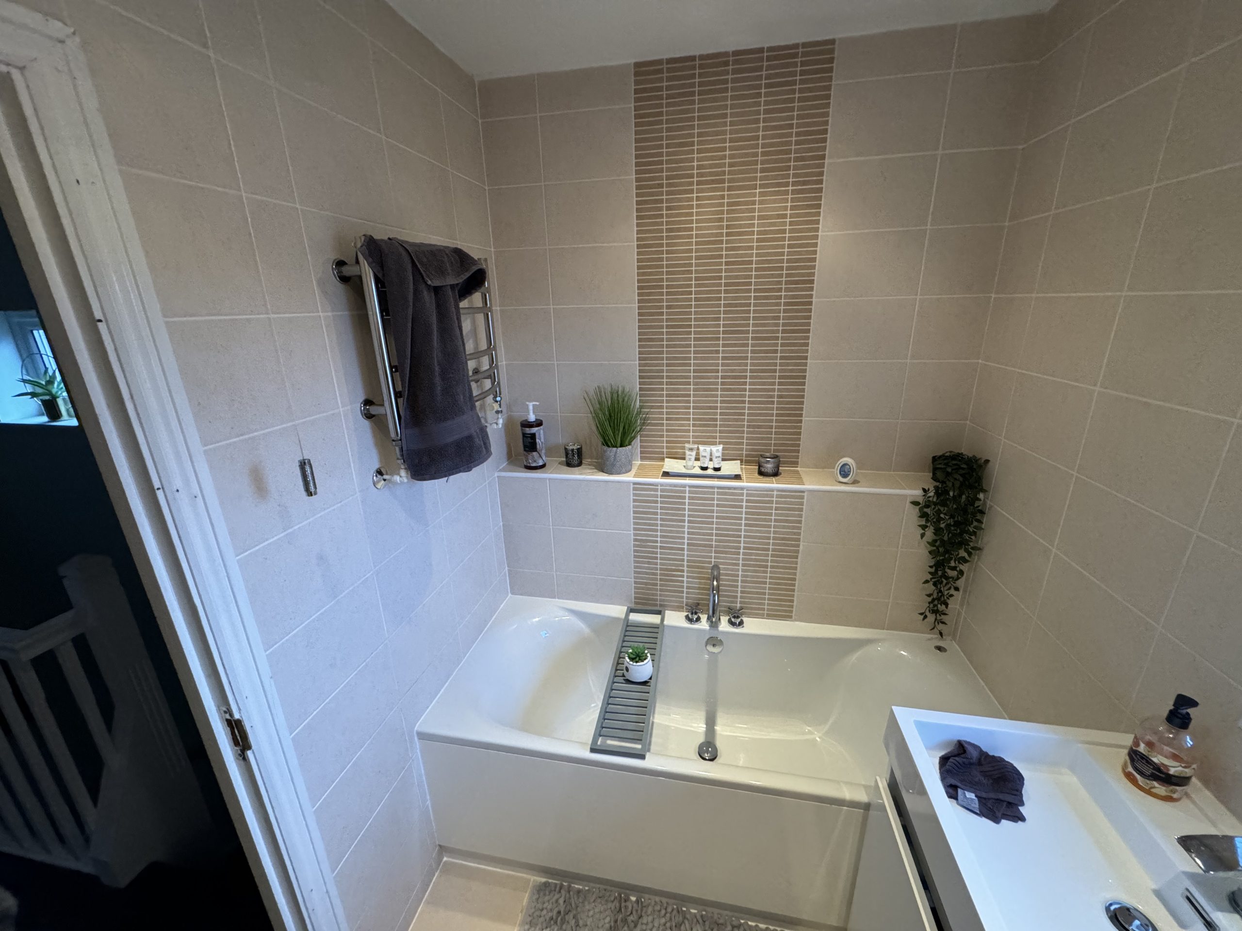 Modern bathroom with beige tiled walls, a white bath, silver fittings, and plants on a shelf—showcasing stylish design ideas perfect for any kitchen and bathroom showroom installation supply. Towels are neatly stacked, with a bath tray over the tub and a tidy sink corner.