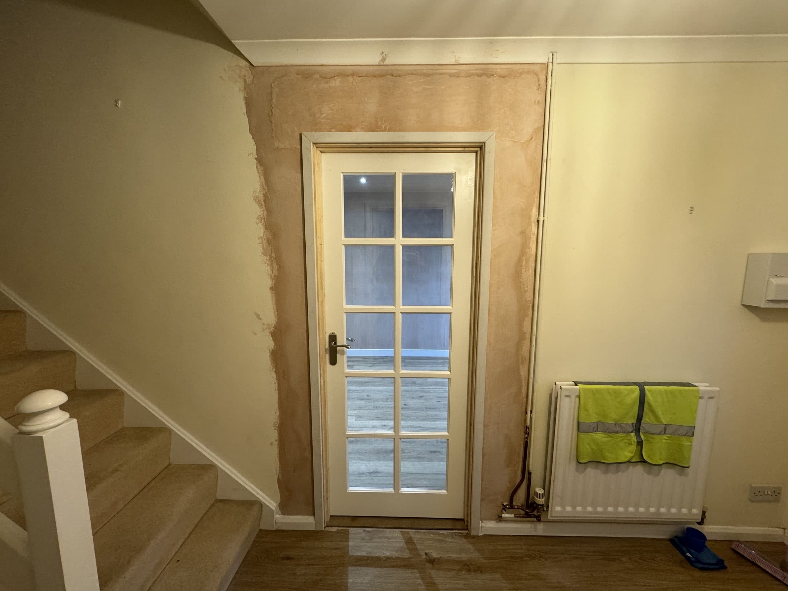 A glass-paned door with unfinished plasterwork around the frame is at the bottom of a staircase in this kitchen and bathroom showroom. A radiator with a high-visibility vest hangs on the wall to the right of the door. The floor is wooden.