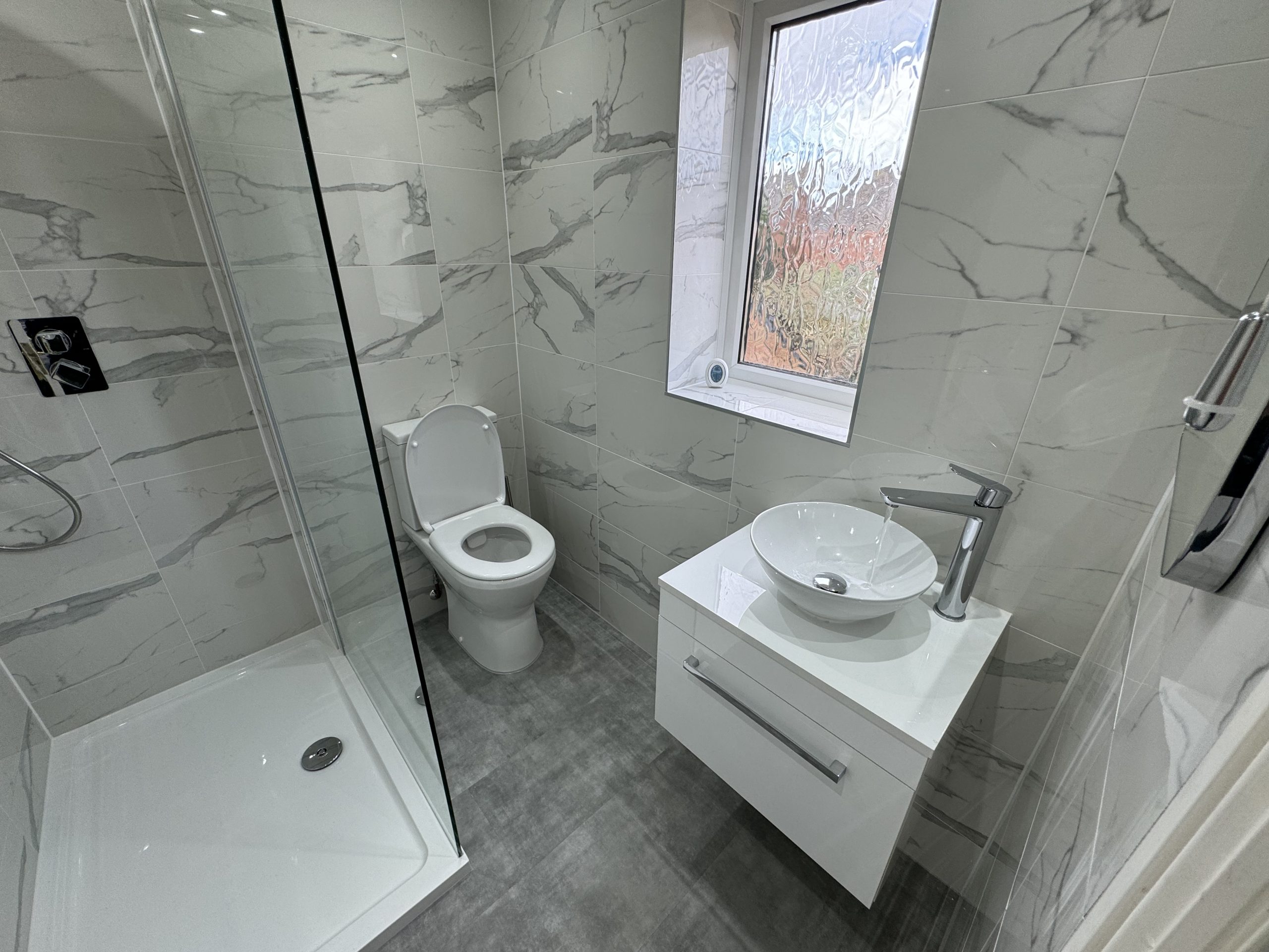 A modern bathroom designed with grey marble tiles, a glass shower enclosure, white toilet, and floating vanity—showcasing premium kitchen and bathroom showroom installation and supply expertise.