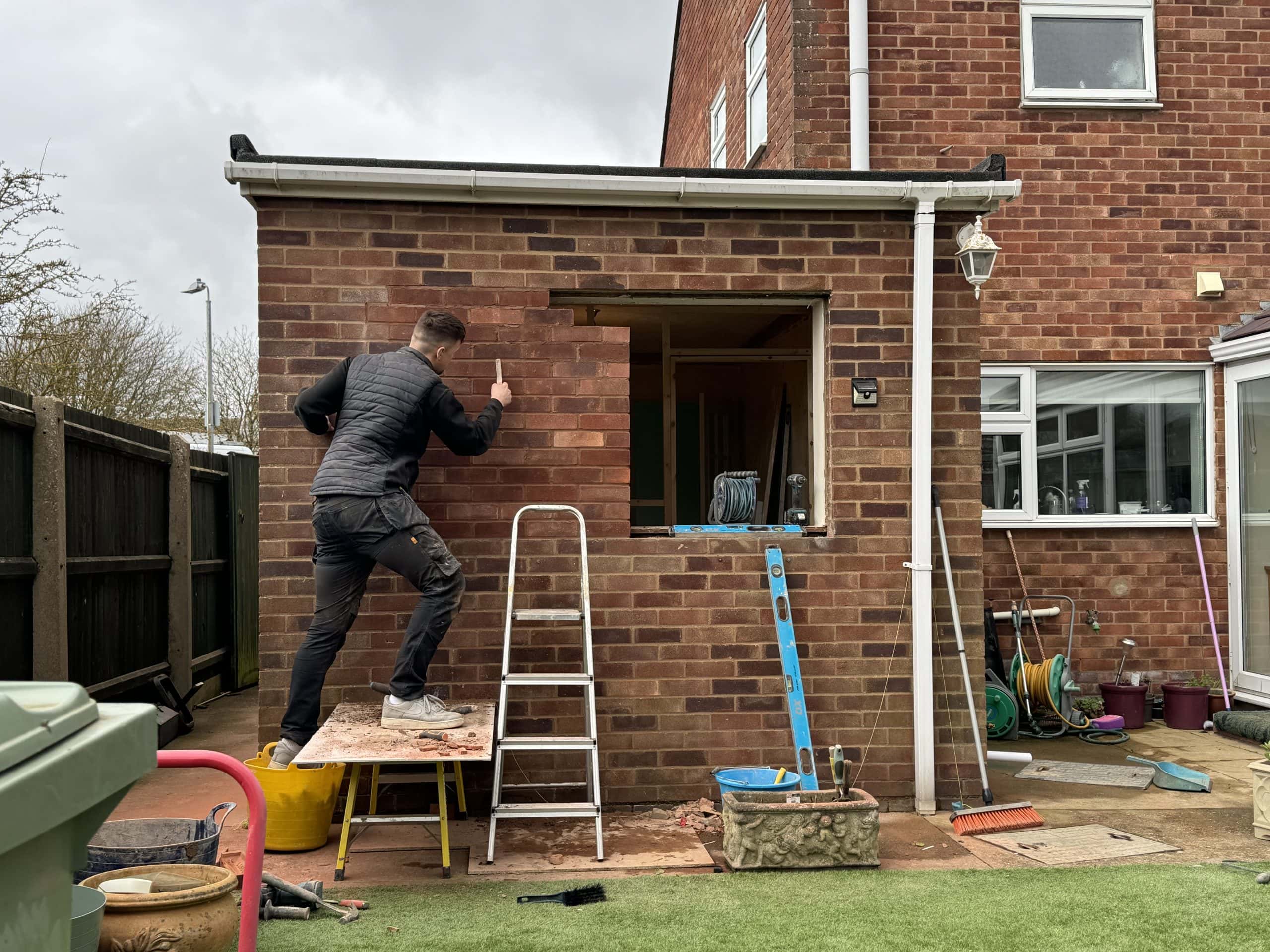 A person stands on a wooden platform beside a ladder, working on the brick wall of a house extension. Tools for kitchen and bathroom showroom installation are scattered around the garden, and an open window reveals the unfinished interior.