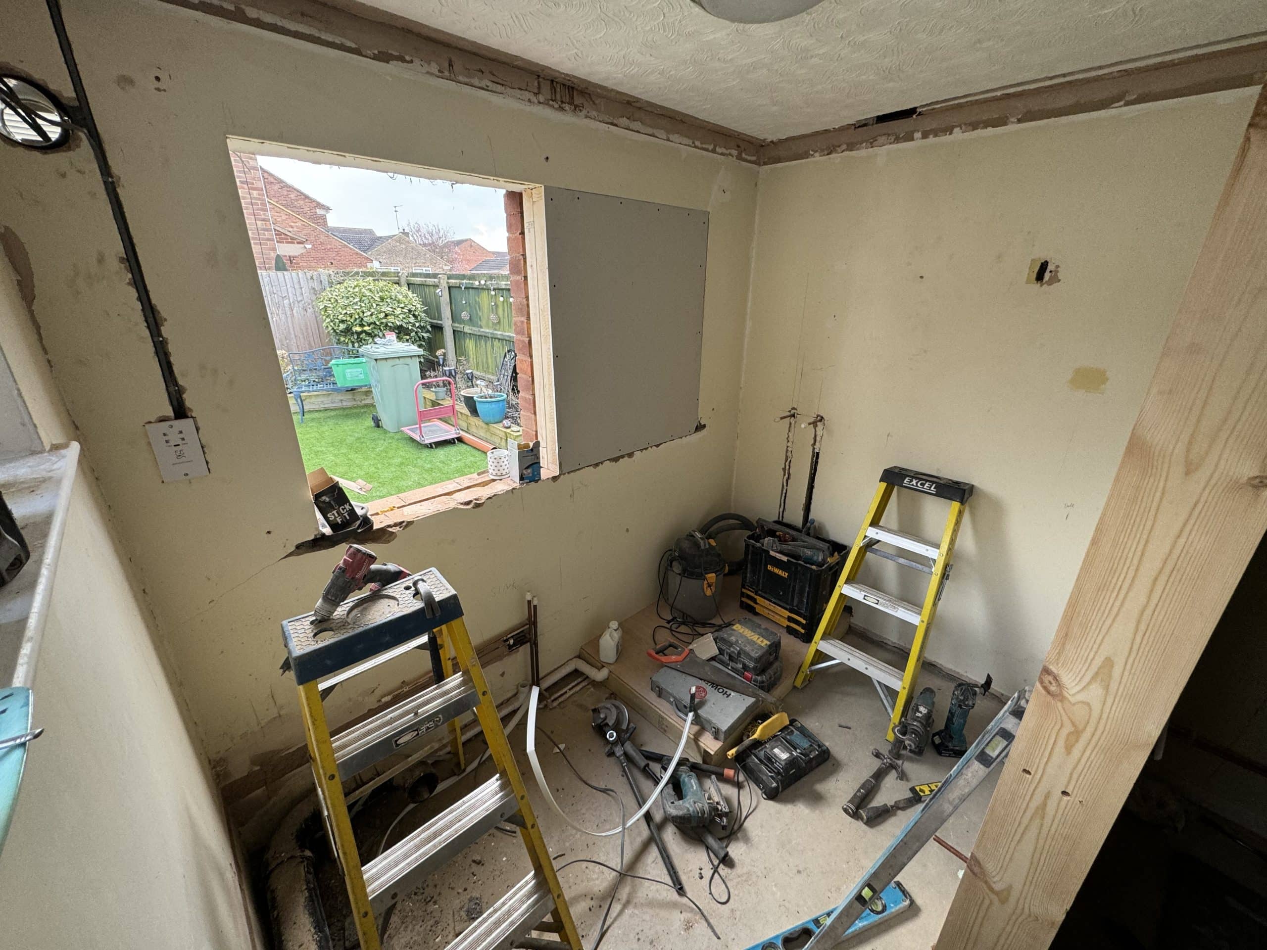 A small room under renovation with tools, ladders, and kitchen installation equipment scattered on the floor. A large hole is cut into one wall, revealing a view of a back garden with grass, plants, and a fence.