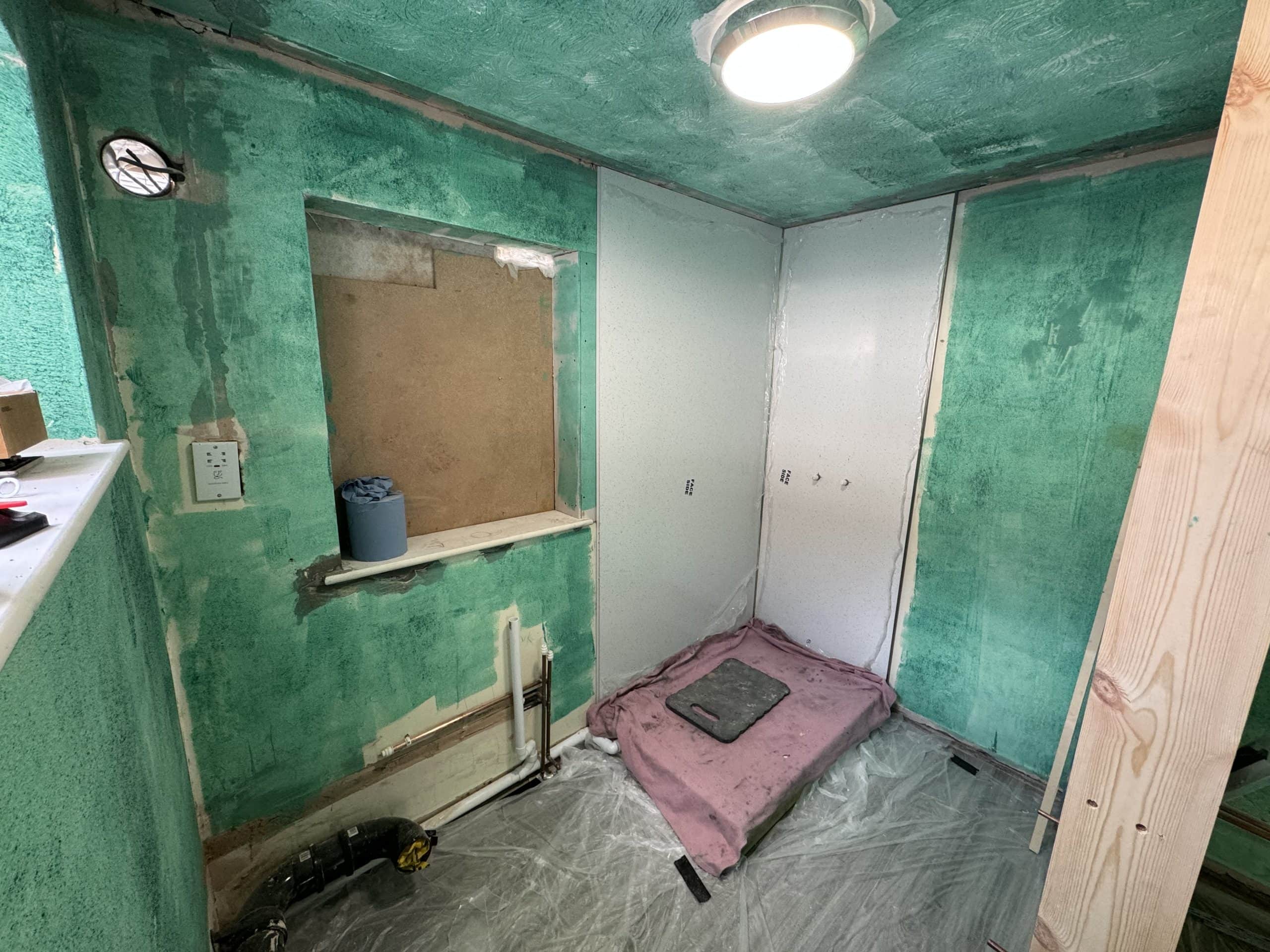A small room undergoing renovation with green waterproofing on the walls, plastic sheeting covering the floor, a recessed window, exposed pipes, and scattered building materials—a glimpse into a future kitchen and bathroom showroom installation. A ceiling light is on.