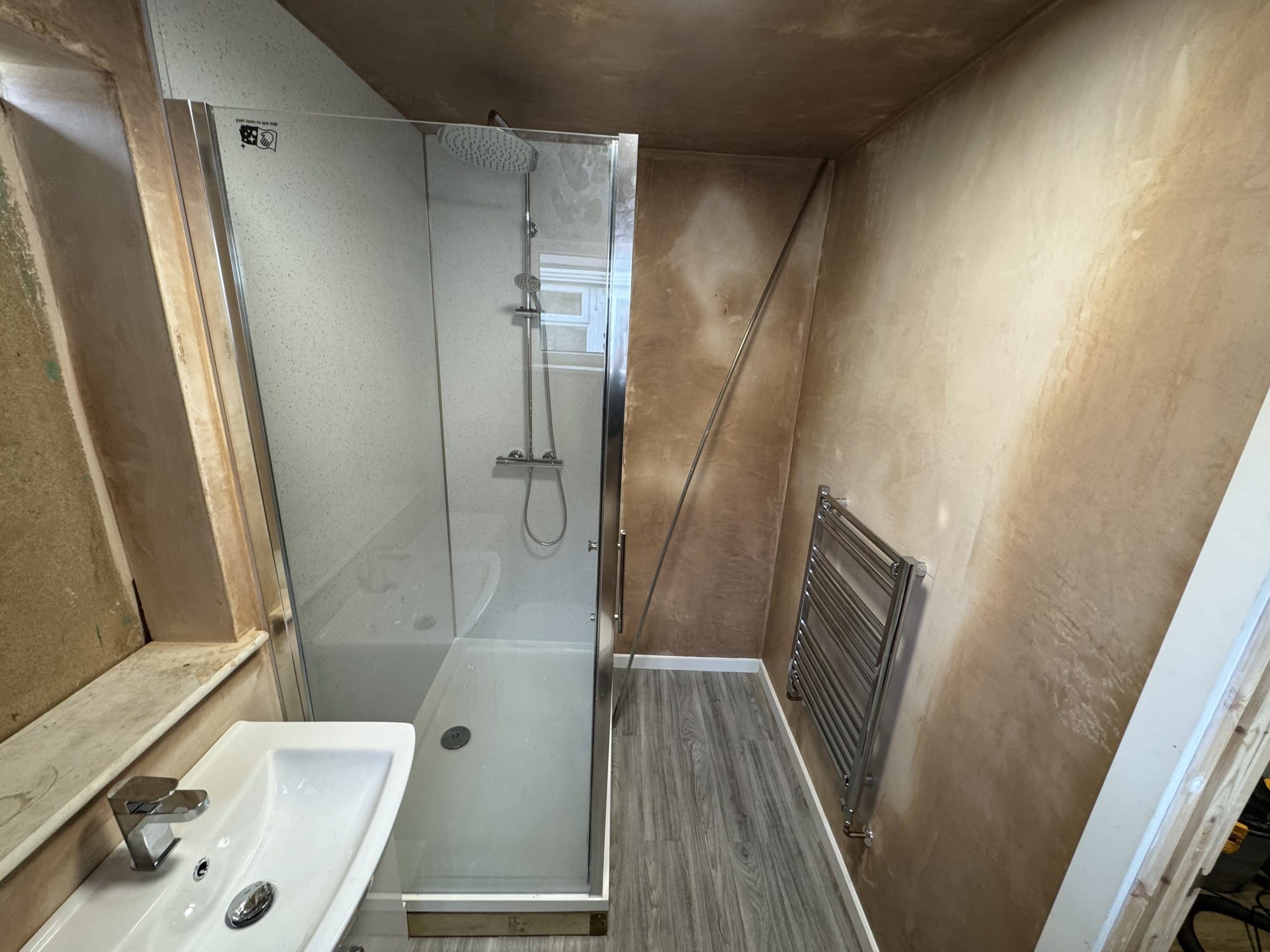 A newly renovated bathroom, designed with showroom-quality features including a glass shower enclosure, rainfall showerhead, silver towel radiator, white basin with chrome tap, and light wood-look flooring. Walls are unfinished plaster.