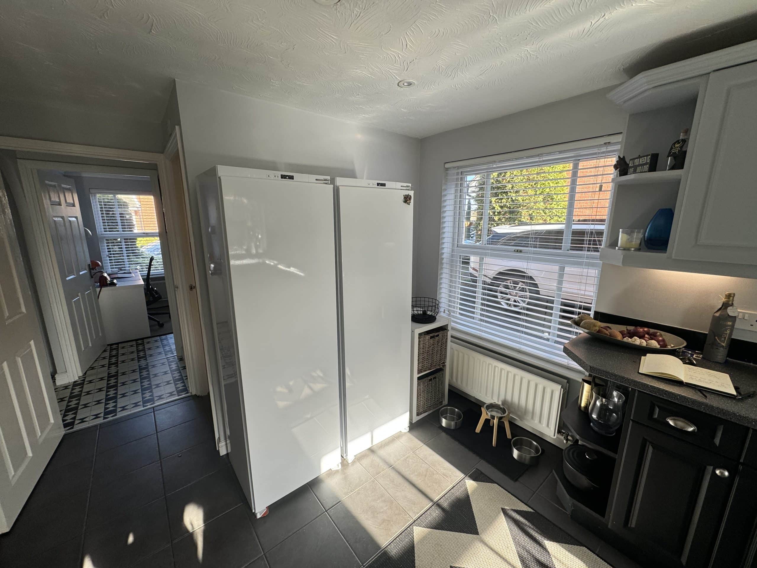 Bright kitchen designed with showroom-inspired touches: two tall white appliances, black worktop with a mixer, fruit bowl, and wine bottle. A window with blinds reveals a white car; food and water bowls rest on the tiled floor.