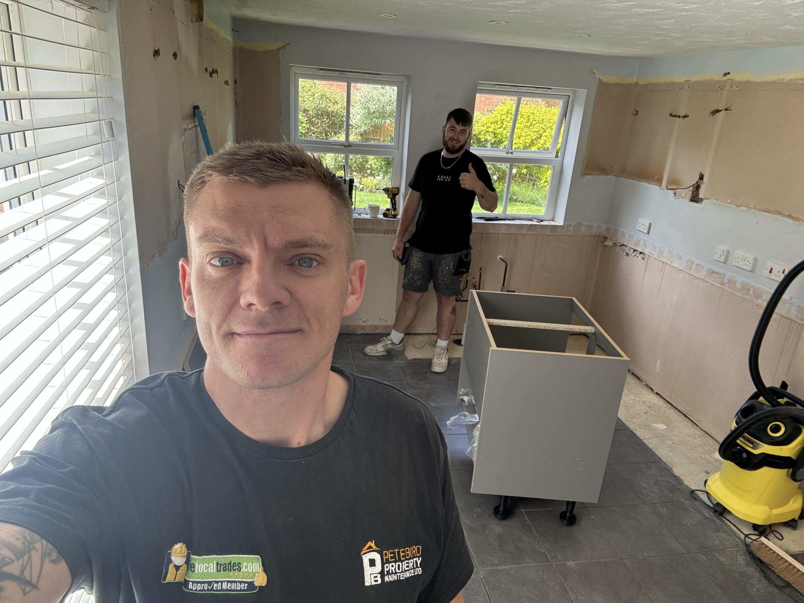 Two men are working in a partially renovated kitchen. One is taking a selfie and smiling, while the other by the window gives a thumbs up. Kitchen showroom installation materials and design tools are visible around them.