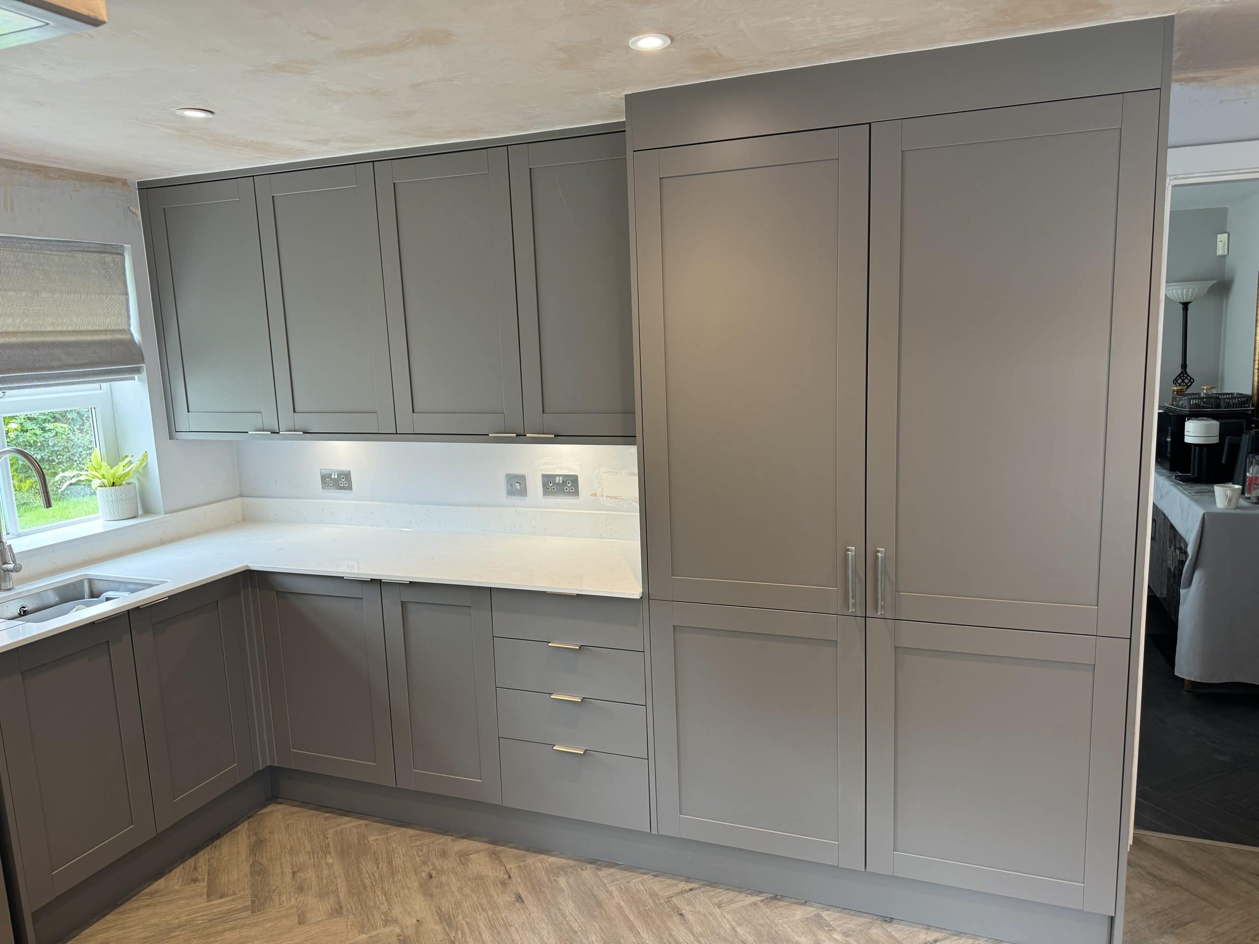 Modern kitchen with matte grey cupboards, white worktops, built-in fridge, and under-cupboard lighting. Ideal for showroom installation or design inspiration, this clean, minimalist space features wood-patterned flooring and a window above the sink.