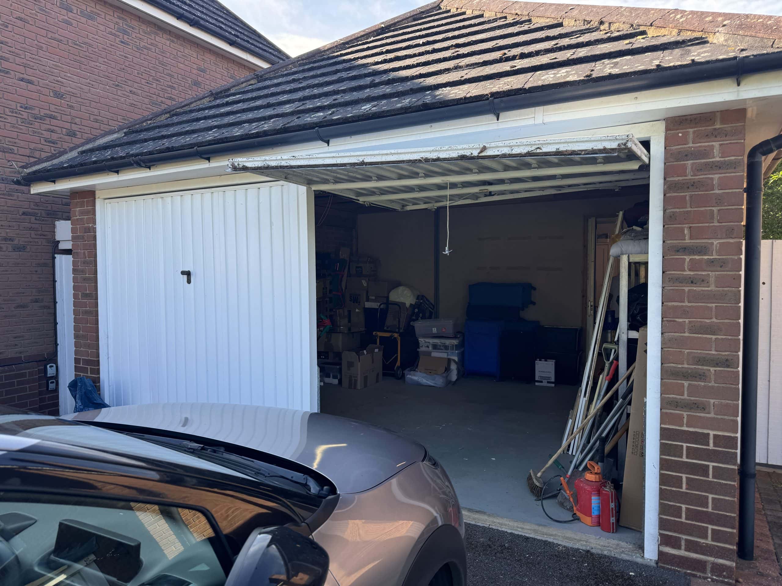 A partially open garage door reveals a cluttered space filled with storage boxes, tools, and equipment—some of which hint at kitchen and bathroom showroom installation projects. A silver car stands parked outside, its bonnet visible in the foreground.