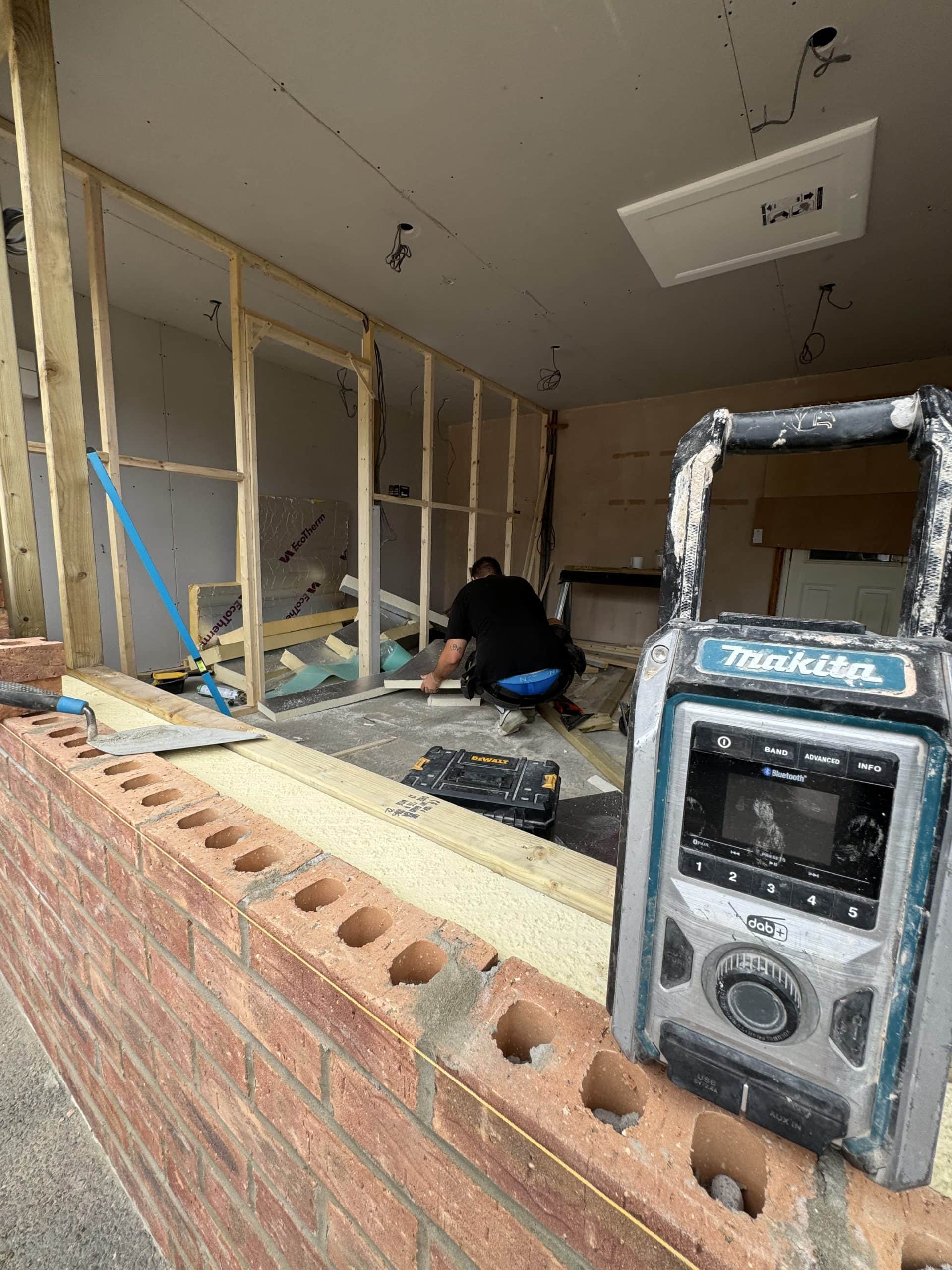 A construction site with a worker kneeling on the floor installing insulation; an unfinished brick wall is in the foreground and a Makita radio sits on the wall. Framing and tools for kitchen and bathroom installation design are visible throughout the scene.