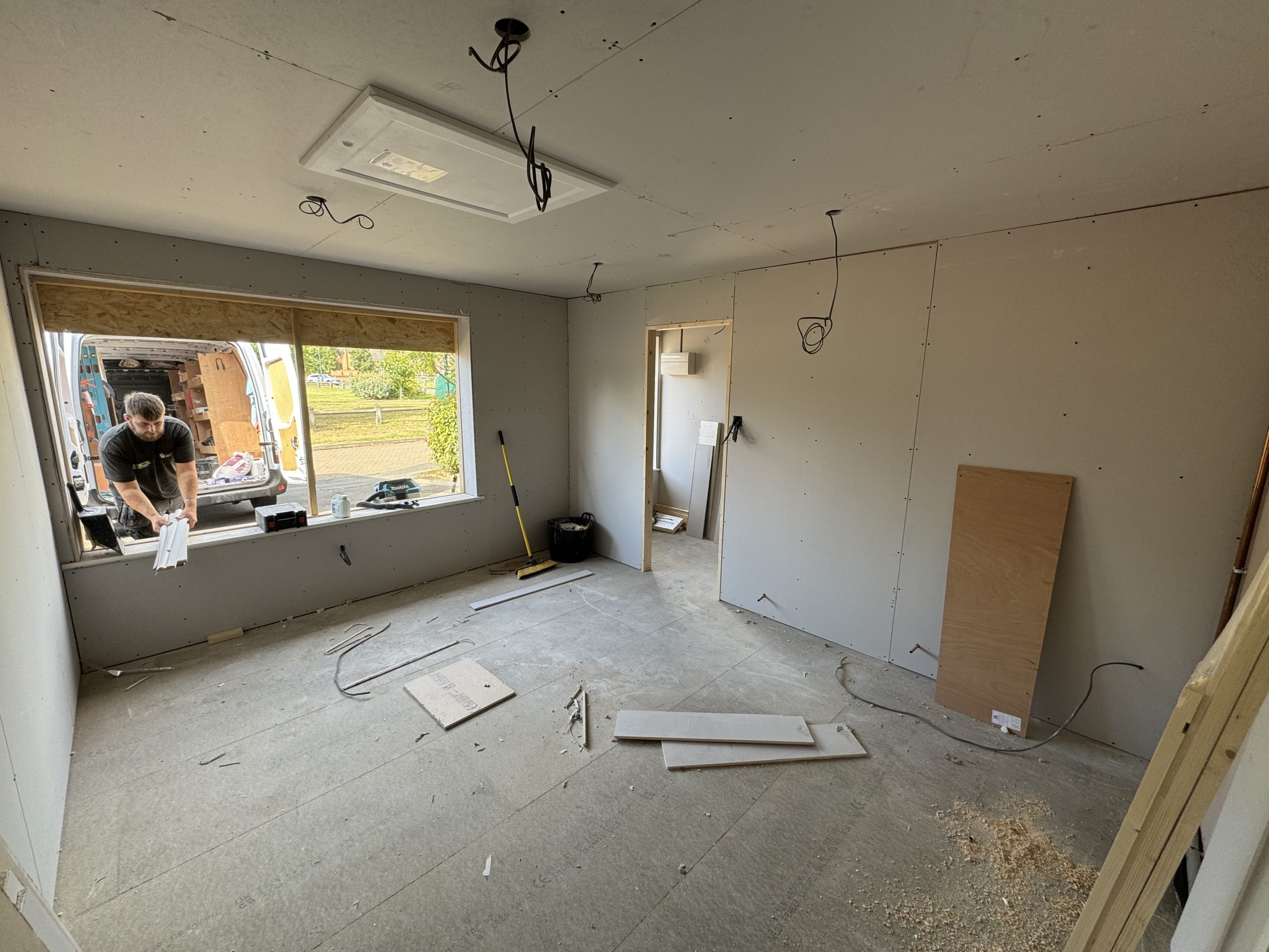 A room under construction with unfinished walls and exposed wiring. A man works near a large open window, surrounded by kitchen and bathroom showroom installation materials and tools scattered on the floor. A van is visible outside.