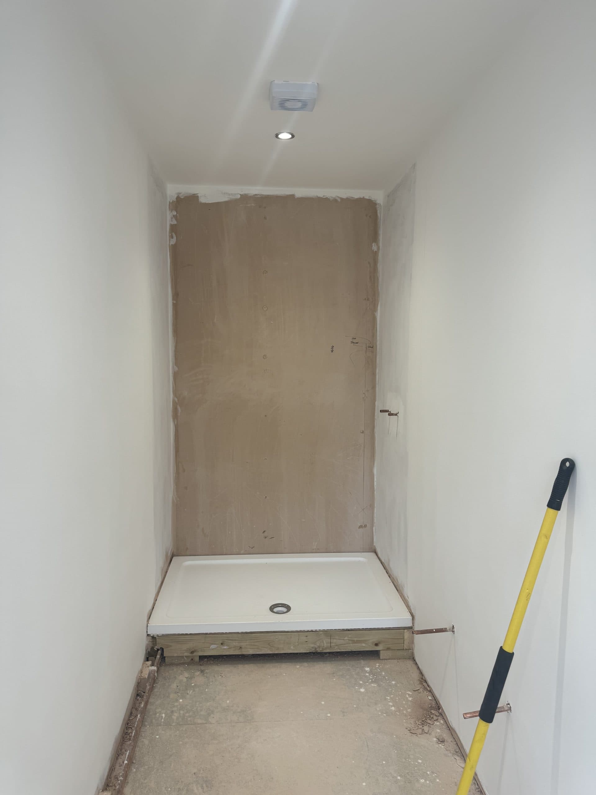 A small, unfinished bathroom with white walls, a bare plaster wall at the end, a white shower tray installed, and a yellow-handled tool leaning against the right wall. Reflects the early stage of a bathroom showroom installation or design project.