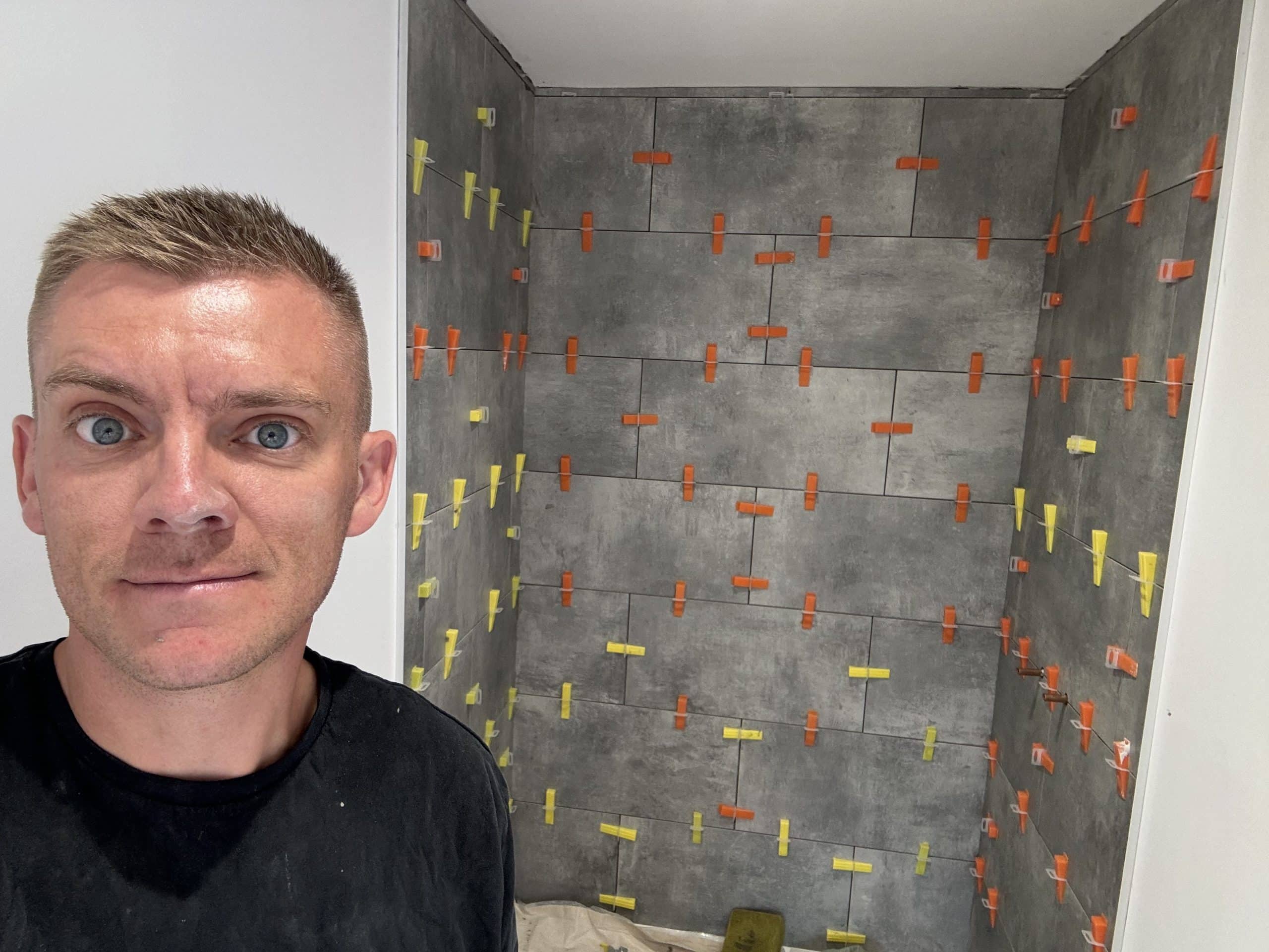 A person stands before a tiled bathroom wall, featuring grey tiles and numerous orange and yellow spacers—showcasing meticulous installation work inspired by modern kitchen and bathroom showroom design.