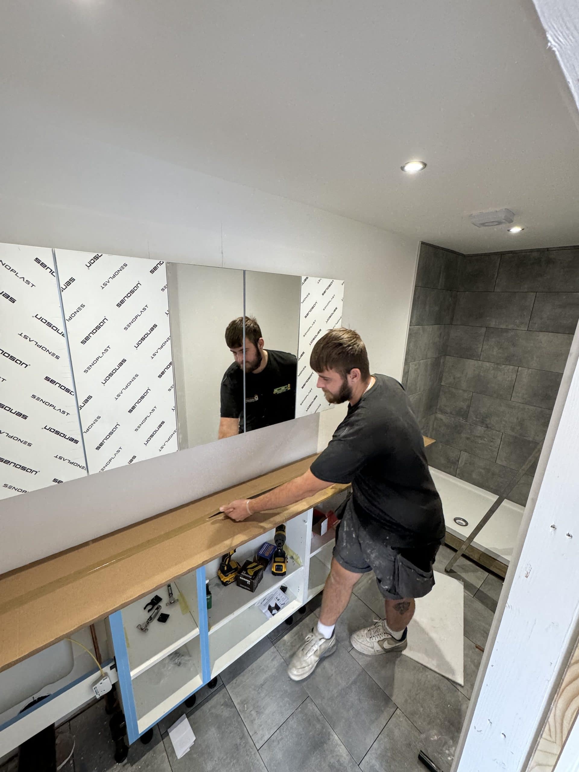 A man in work clothes measures and cuts a board on top of a bathroom vanity unit, surrounded by tools. The space showcases a kitchen and bathroom showroom design, with grey tiled walls, a shower tray, and large mirrors above the vanity unit.