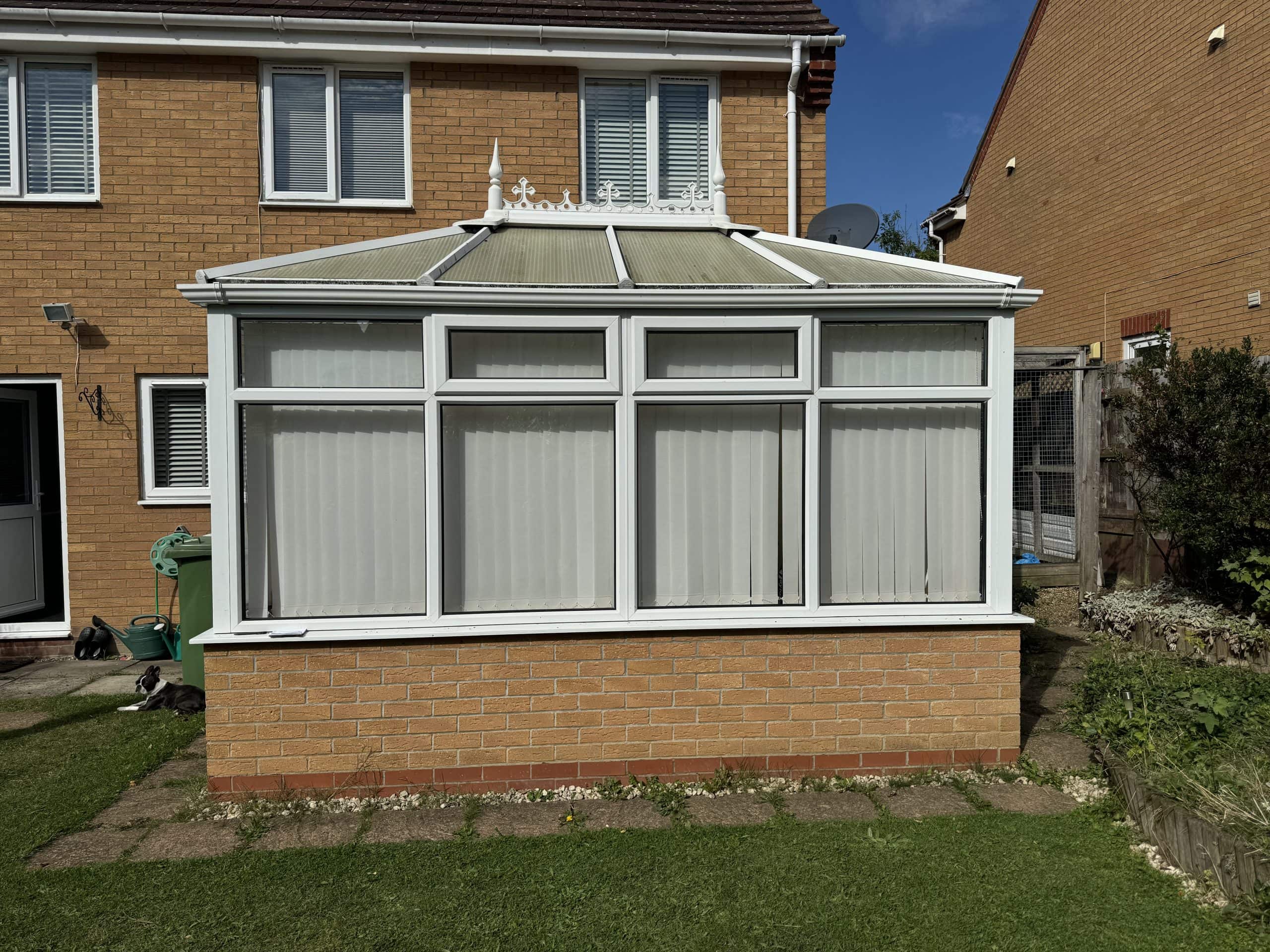 A white conservatory with glass roof and vertical blinds attached to a brick house, with kitchen and bathroom showroom design inspiration, and a small lawn and garden area in the foreground under a blue sky.