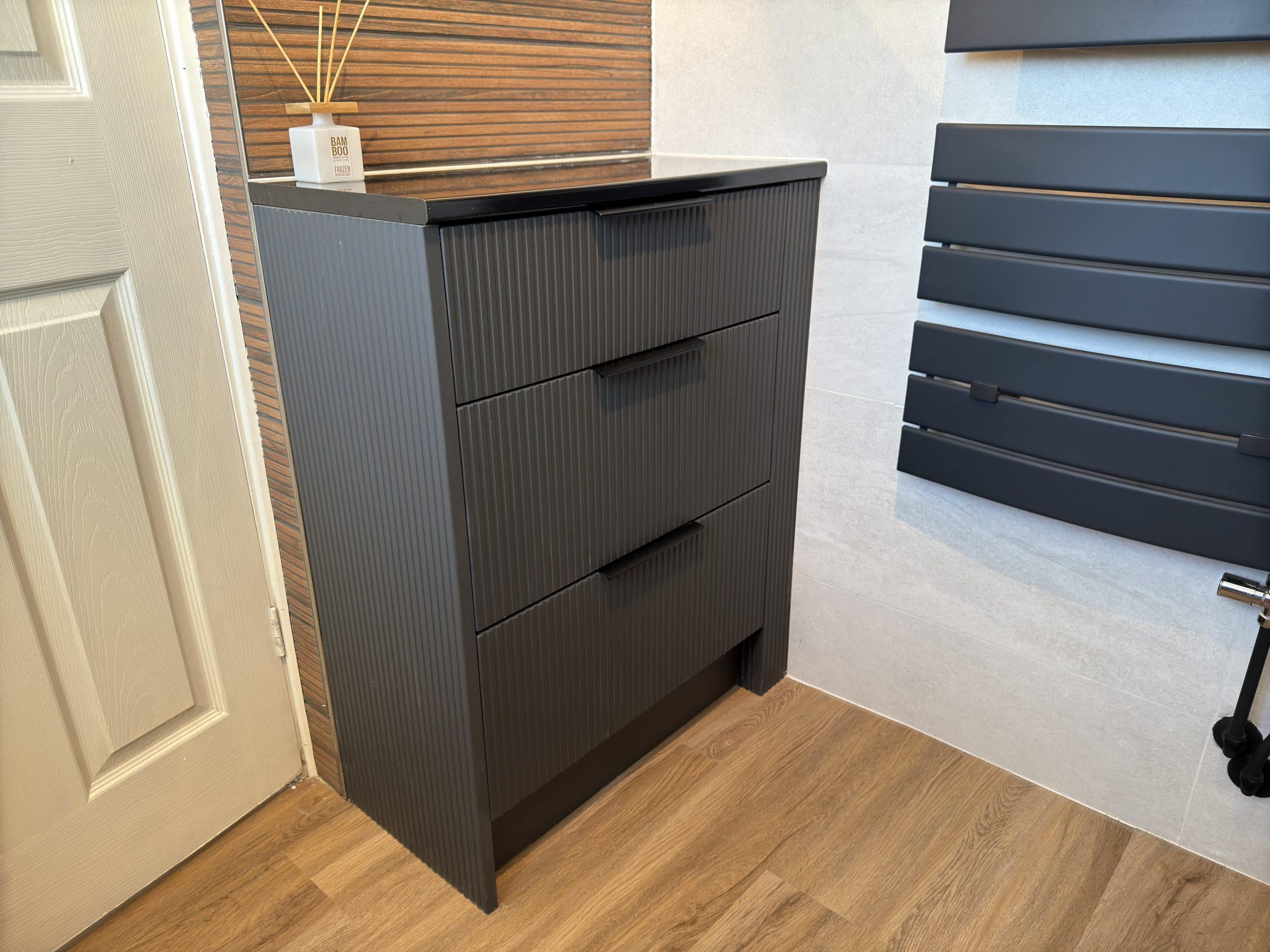 A modern, dark grey chest of drawers stands on a wooden floor beside a white door and wood-panelled wall—ideal for kitchen and bathroom showroom design. Decorative reed diffusers sit on top of the chest.