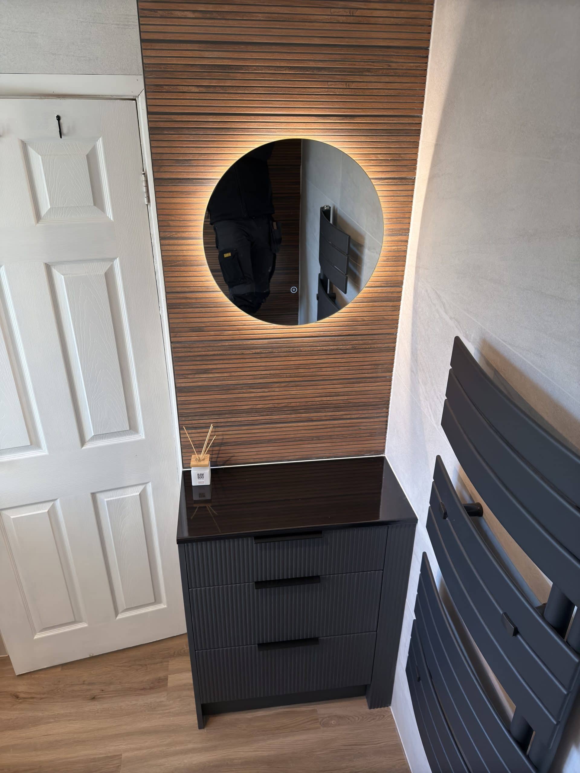 A modern hallway corner designed with a black chest of drawers, round backlit mirror on a wooden slat wall, sleek kitchen and bathroom showroom installation elements, a white reed diffuser, white door, and dark grey wall-mounted radiator panels.