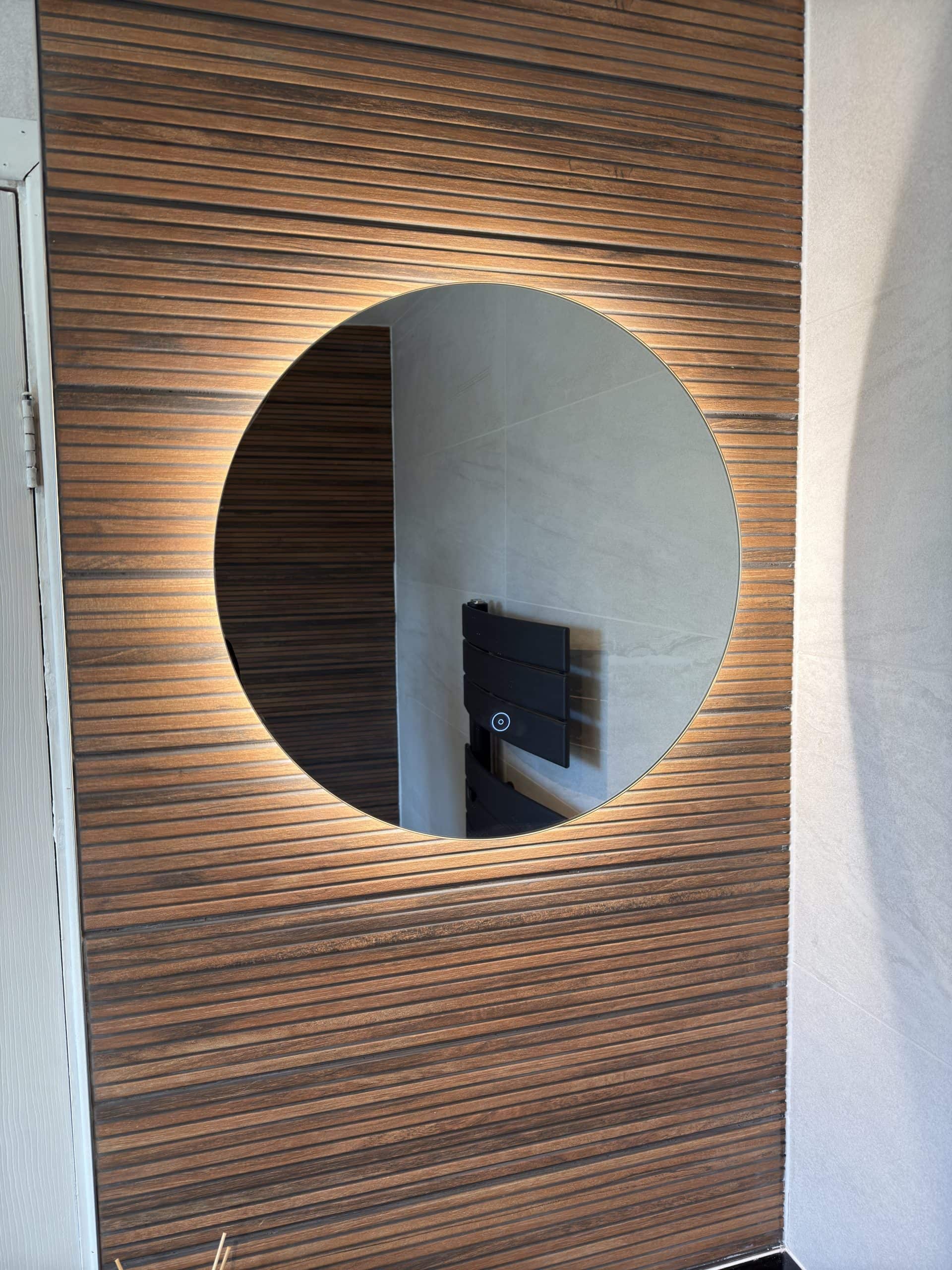 A large round mirror with a backlight is mounted on a wall with horizontal wooden panelling, reflecting a modern interior featured in our kitchen and bathroom showroom installation, with light-coloured walls and a black towel radiator.
