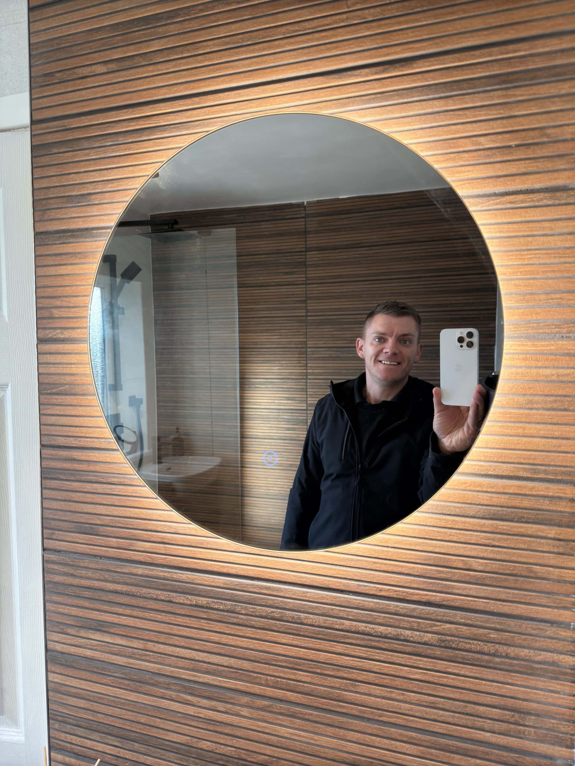 A person is taking a selfie in a round, illuminated mirror mounted on a wood-panelled bathroom wall. Smiling and holding a phone, they showcase the stylish design found in top kitchen and bathroom showroom installations.