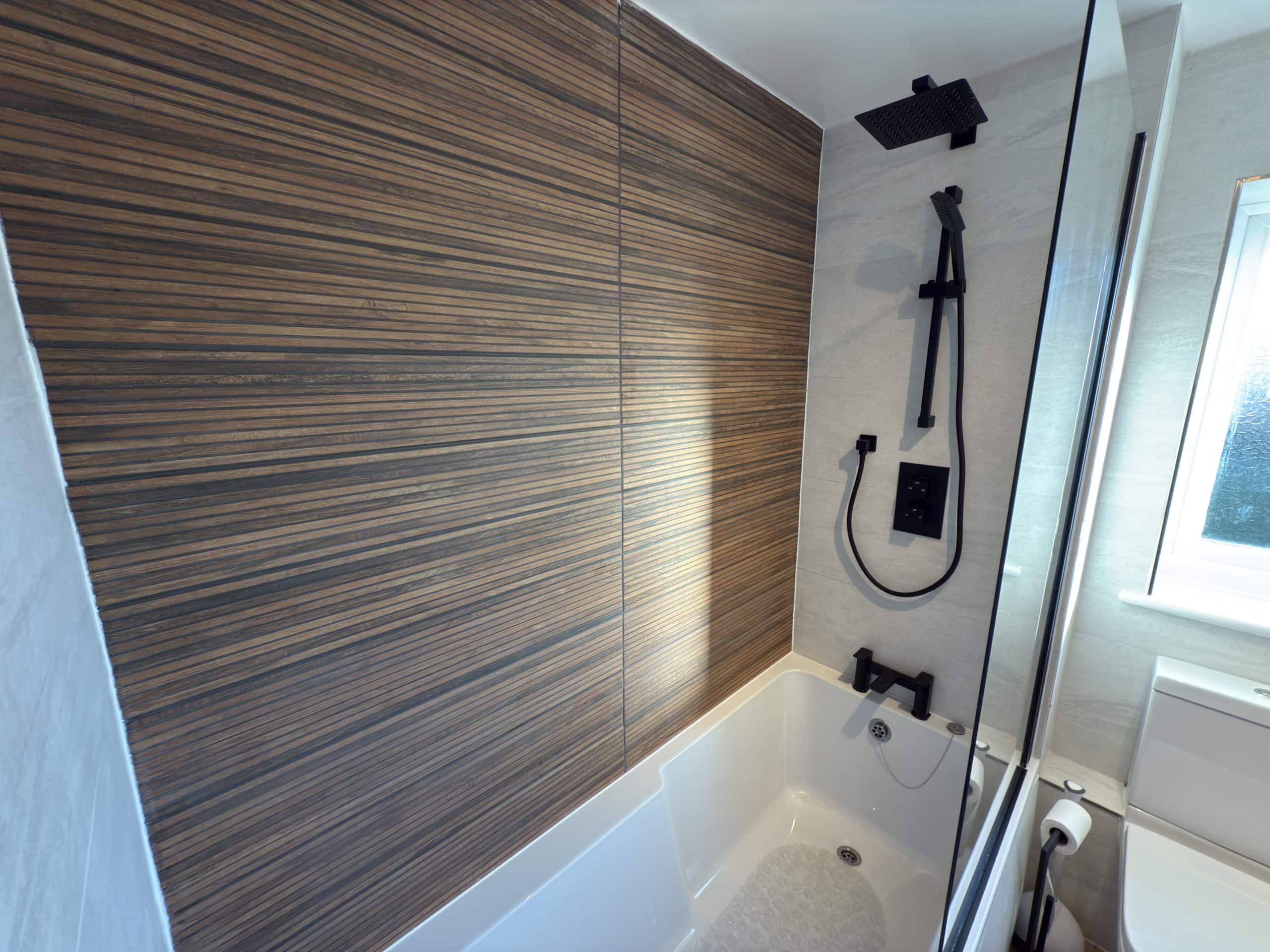 Modern bathroom with a bath, glass shower screen, black rainfall shower head and fittings, and a wood-patterned wall panel next to a window—perfect inspiration from a top kitchen and bathroom showroom installation supply design.