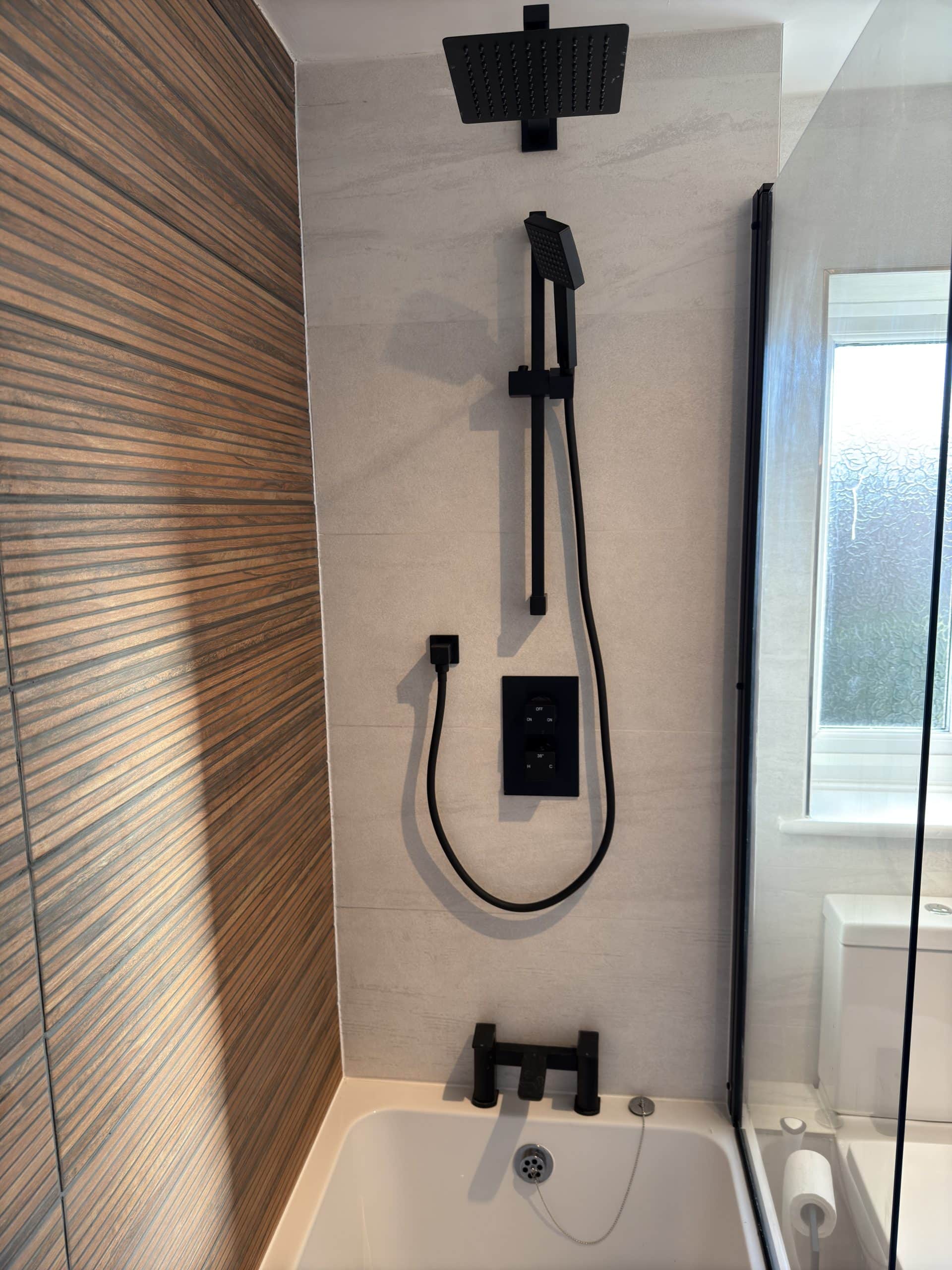 Modern bathroom shower with matt black fittings, including an overhead rainfall shower, handheld shower head, and tap installed above a white bath. Inspired by kitchen and bathroom showroom design, it features neutral tiles and a glass screen near a window.
