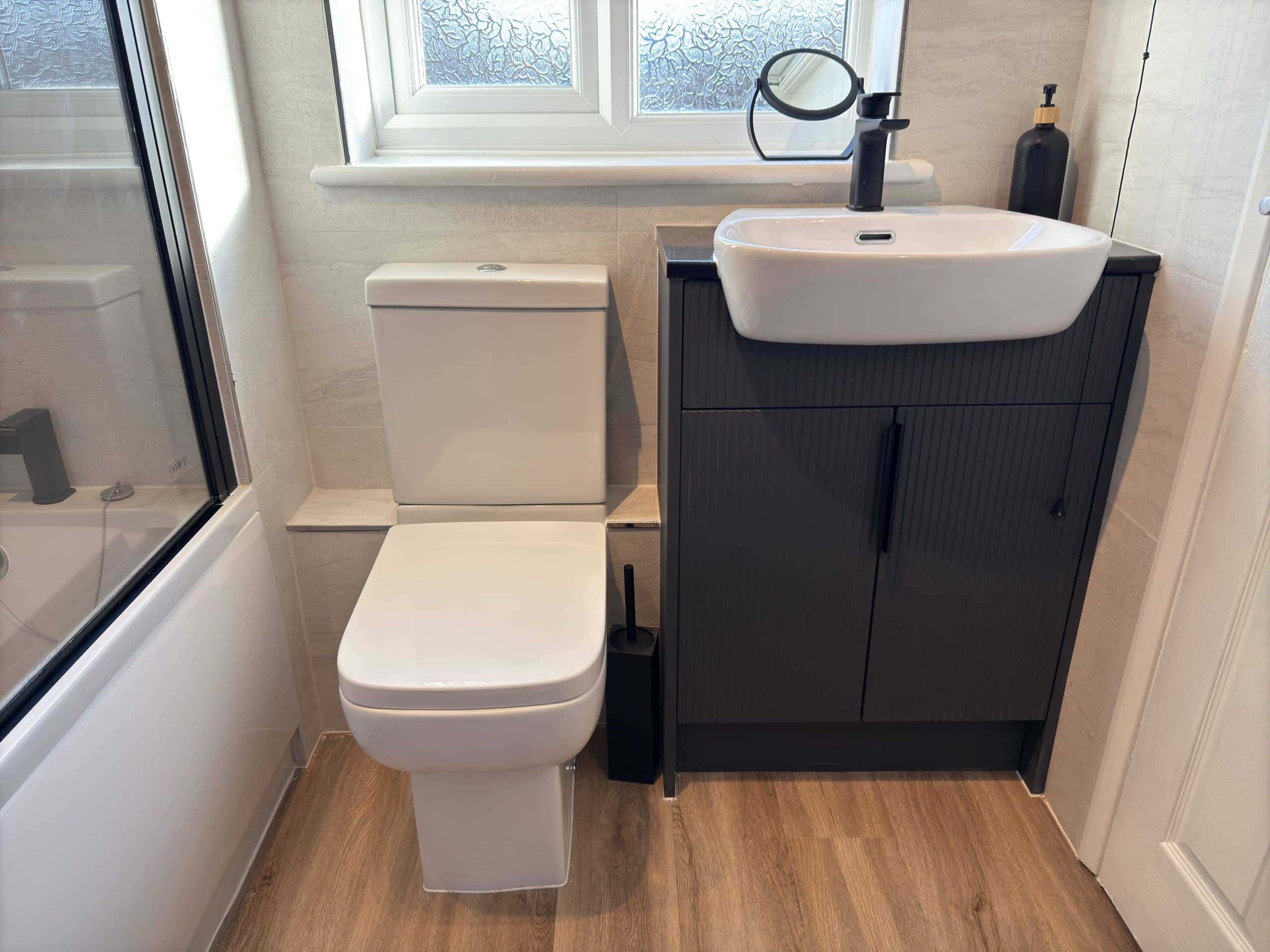 A modern bathroom with a white toilet and black floor brush beside it, next to a black vanity unit with a white washbasin—showcasing sleek kitchen and bathroom showroom design—plus a soap dispenser, round mirror, and wooden floor.