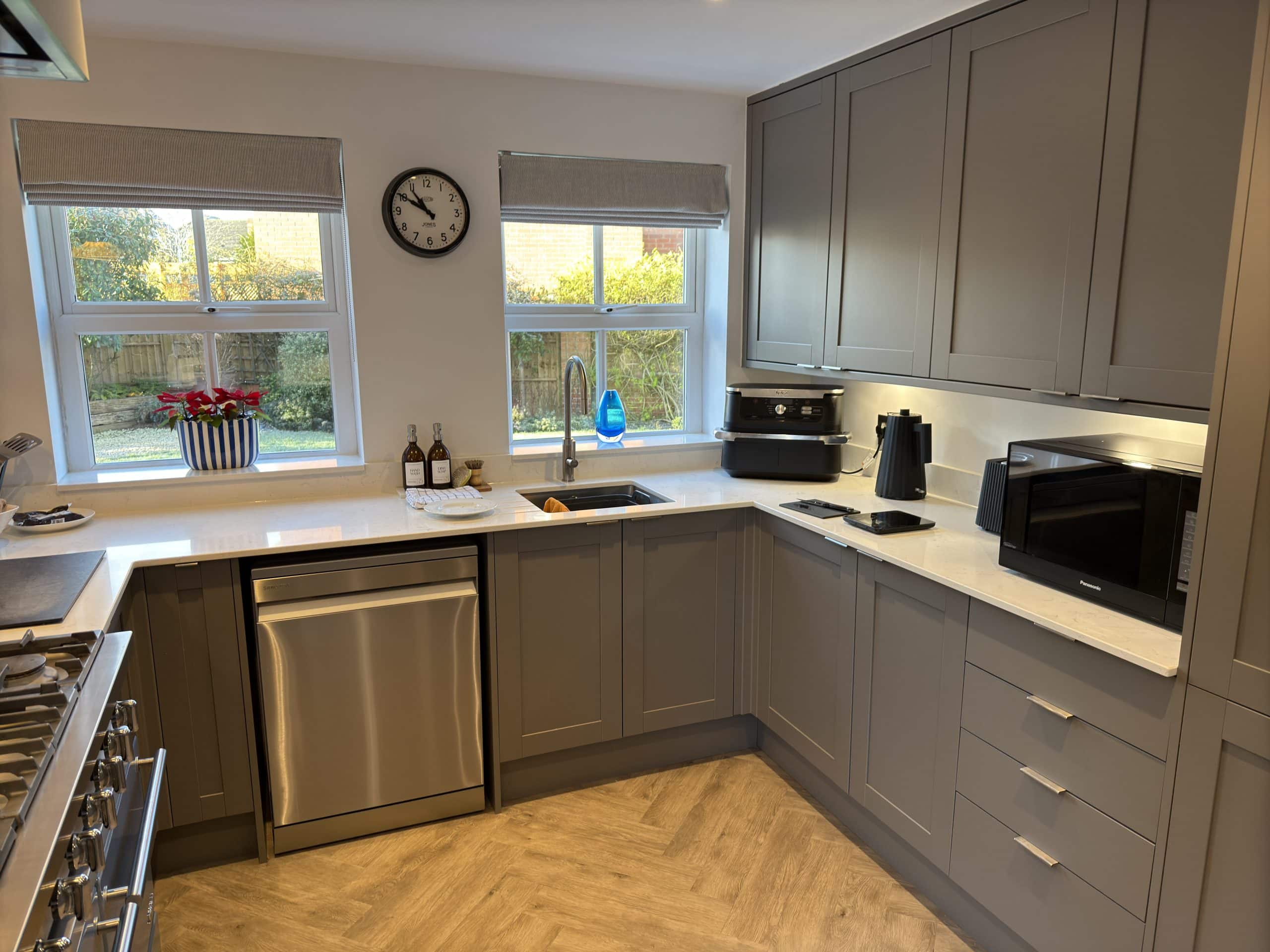 Modern kitchen and bathroom showroom-inspired design with grey cupboards, white worktops, and stainless steel appliances. Natural light streams through two windows adorned with bottles and a vase of red flowers.