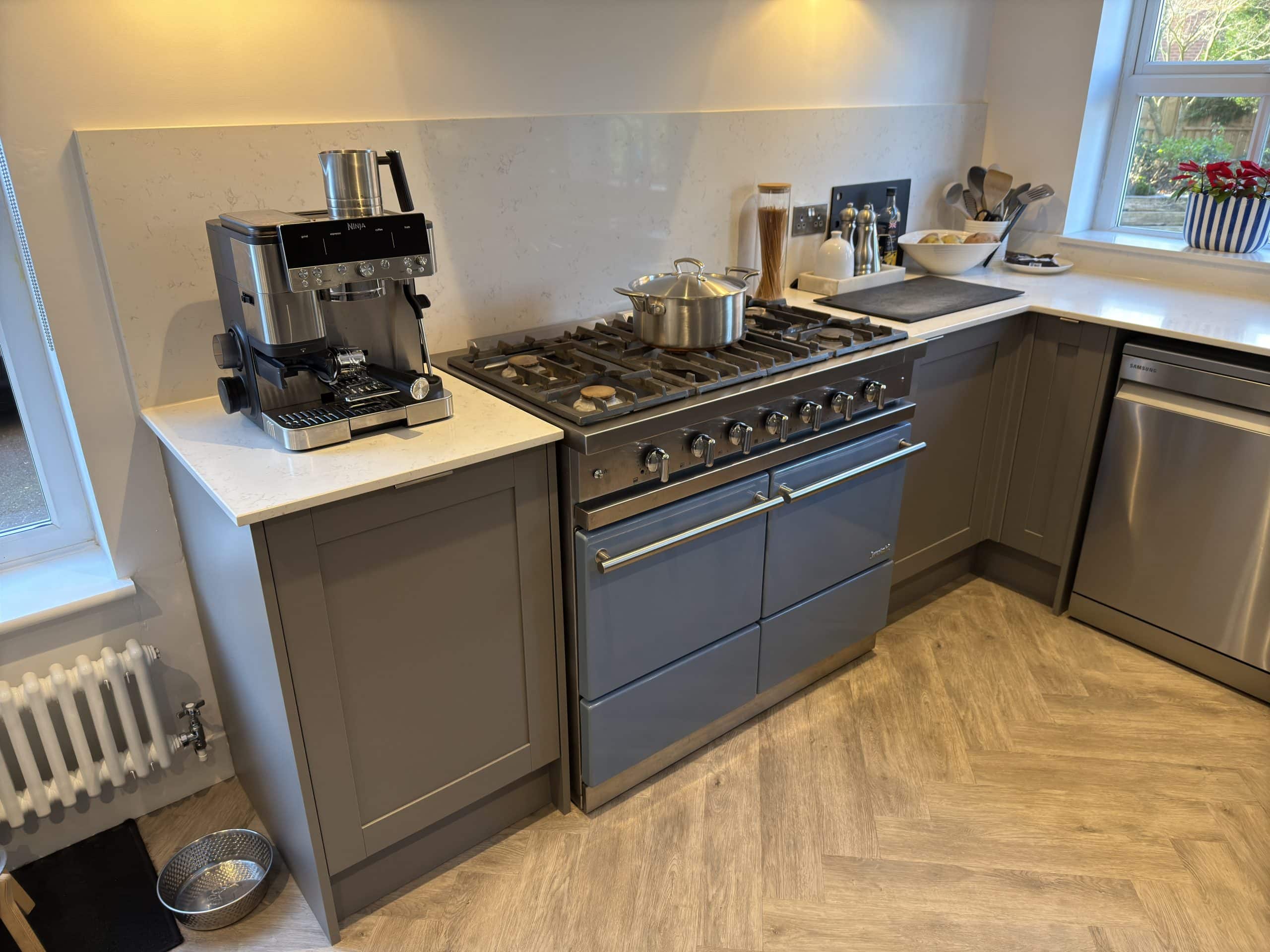 A modern kitchen and bathroom showroom design features grey cupboards, a stainless steel gas cooker with a pot, a coffee machine on the worktop, utensils, a sink, and a bowl of fruit near a window with a potted plant.