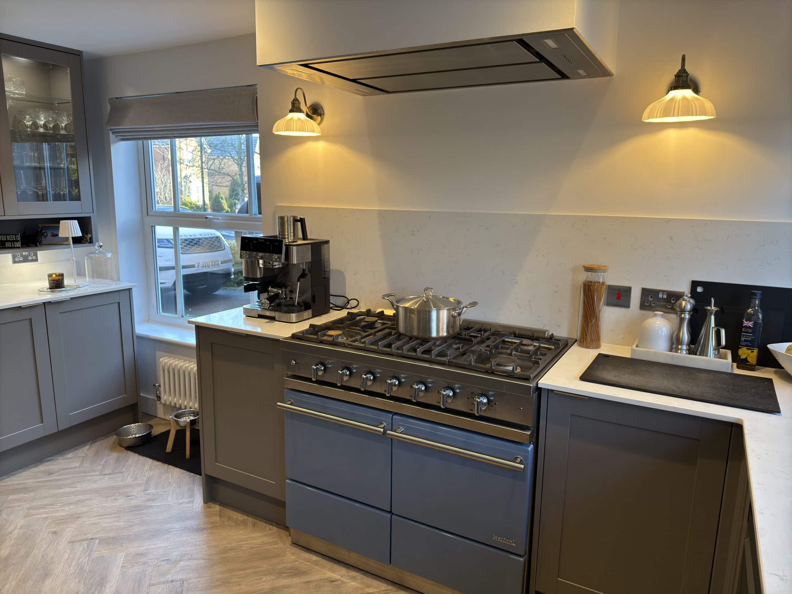 Modern kitchen and bathroom showroom with grey cupboards, a large cooker with saucepans, coffee machine, and utensils on the worktop. Two wall lights illuminate the space, while a window lets in natural light and showcases greenery outside.