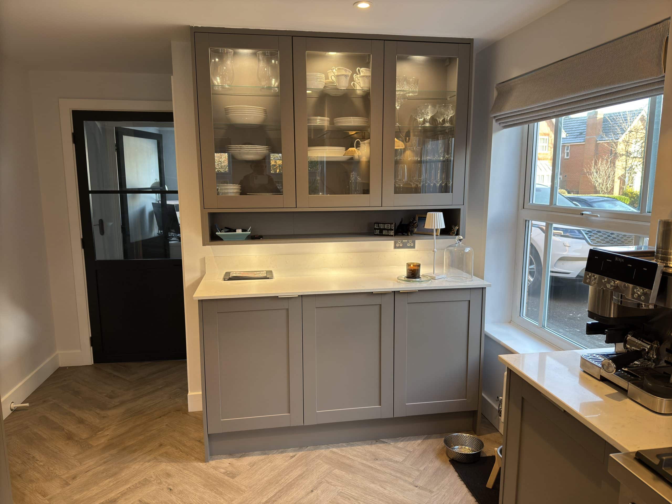 A modern kitchen corner with grey cabinets, glass-fronted upper cupboards displaying crockery, and a white worktop showcases sleek design inspired by kitchen and bathroom showroom installations. A coffee machine sits beside a window, with a dog bowl on the floor.