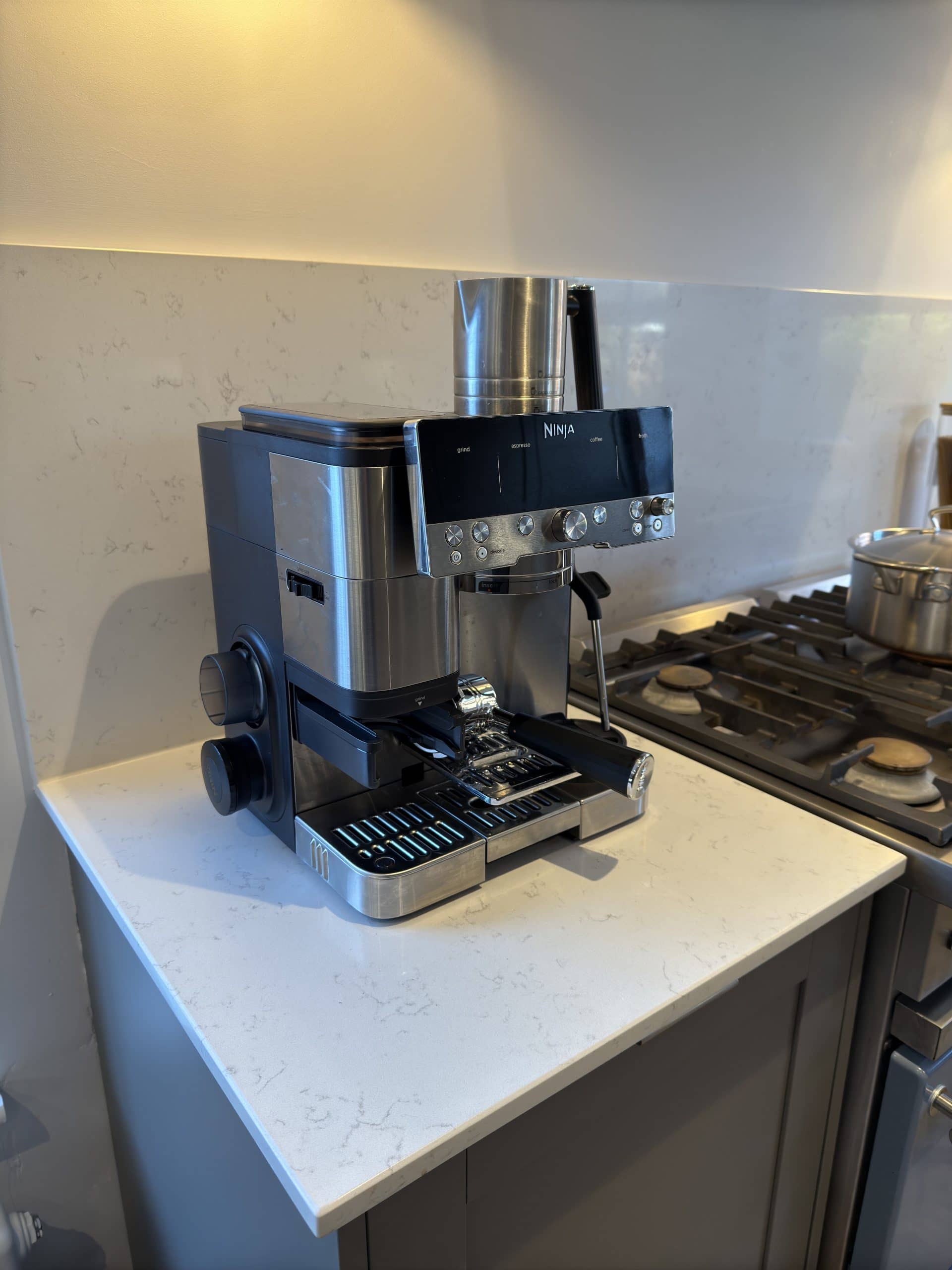 A stainless steel Ninja espresso machine sits on a white countertop next to a gas cooker, showcasing sleek design perfect for any kitchen and bathroom showroom installation, with a portafilter attached and a frothing wand on the side.