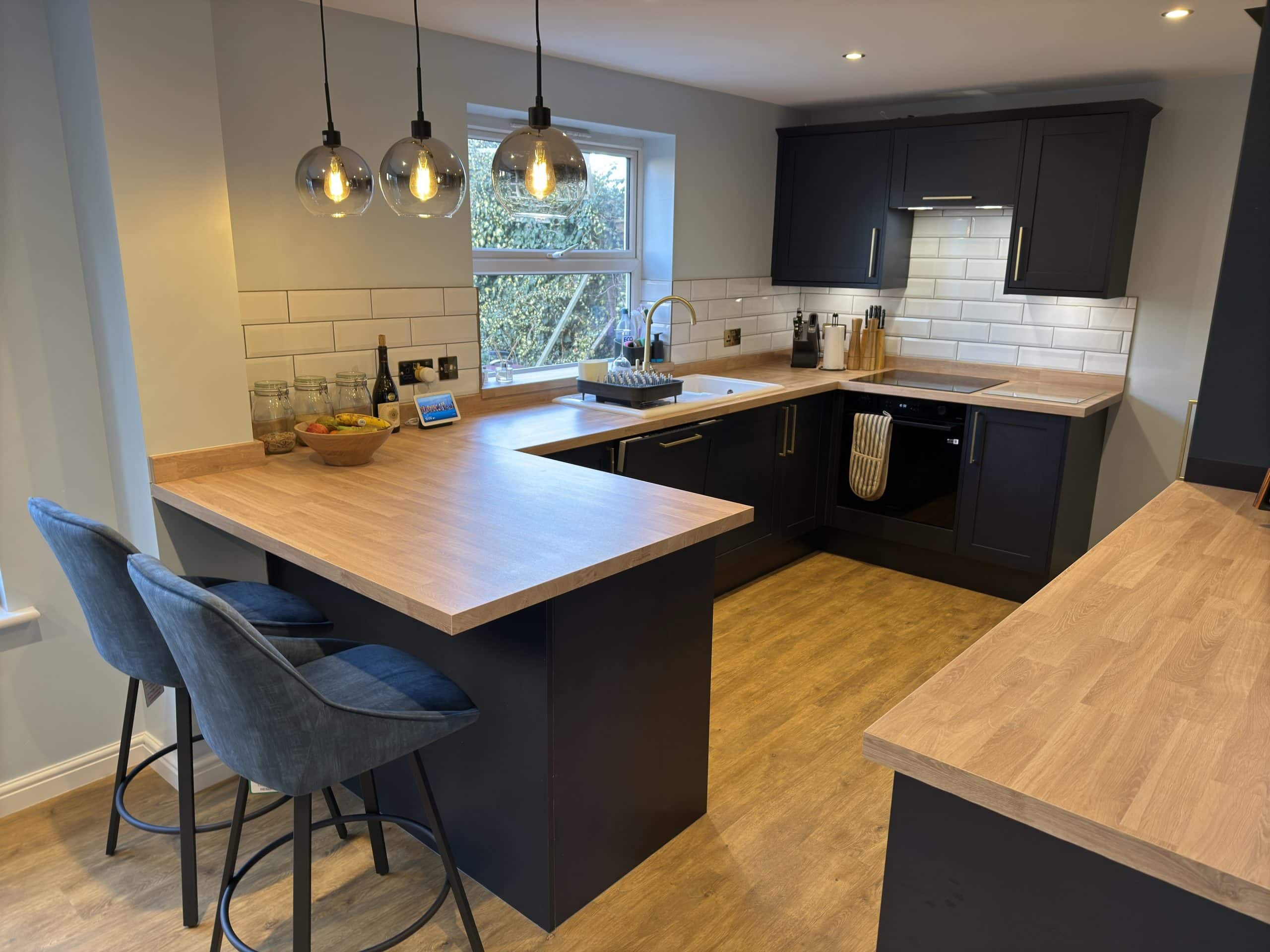Modern kitchen designed with black cupboards, light wood worktops, and a white metro tile splashback. Two grey barstools sit at an island under pendant lights. Ideal for a kitchen and bathroom showroom installation. Natural light streams in through the window.