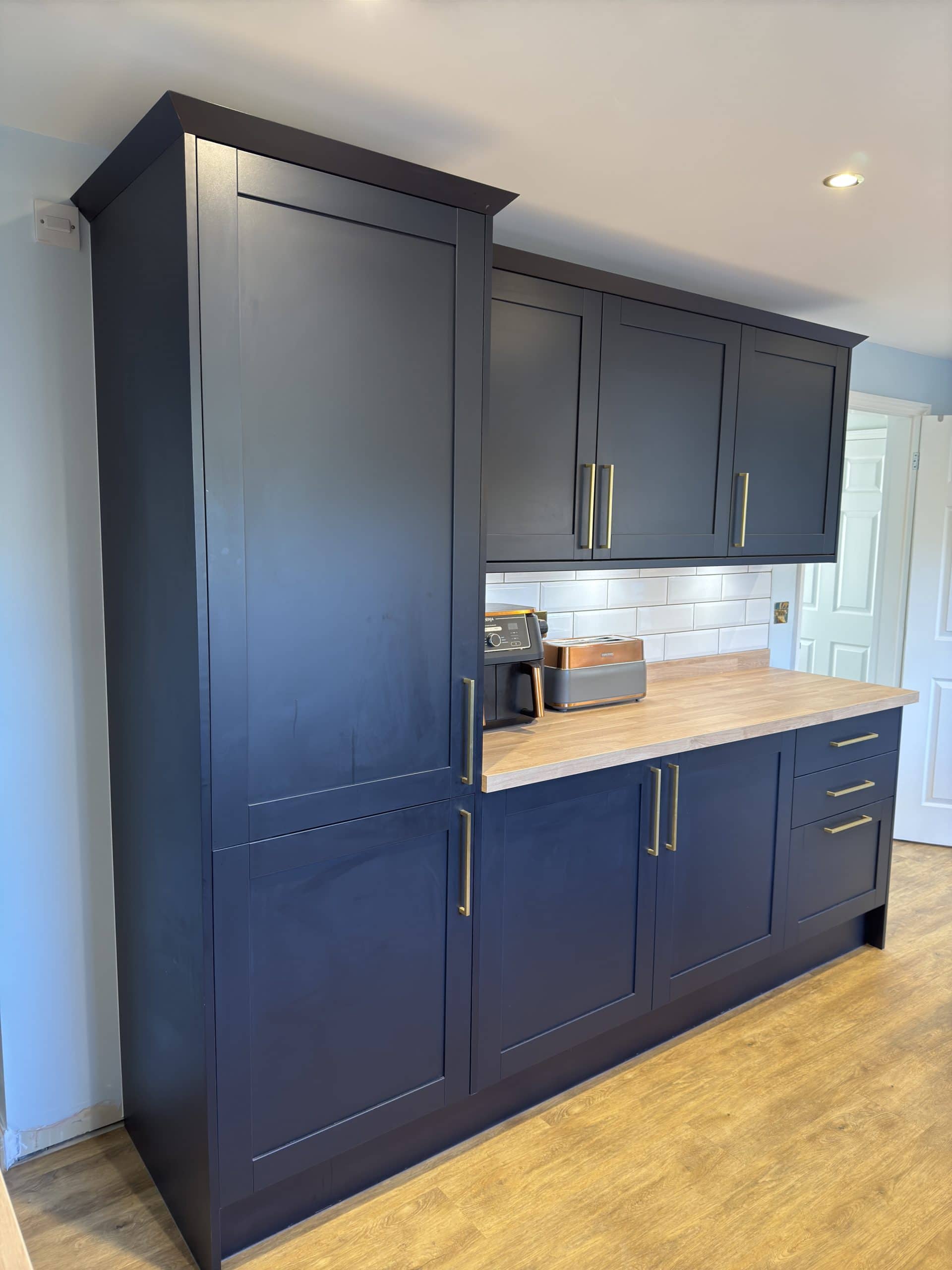 A modern kitchen with dark blue cupboards, gold handles, a light wood worktop, and wood-effect flooring showcases refined design. Small appliances sit on the counter—perfect inspiration for your next kitchen and bathroom showroom installation.