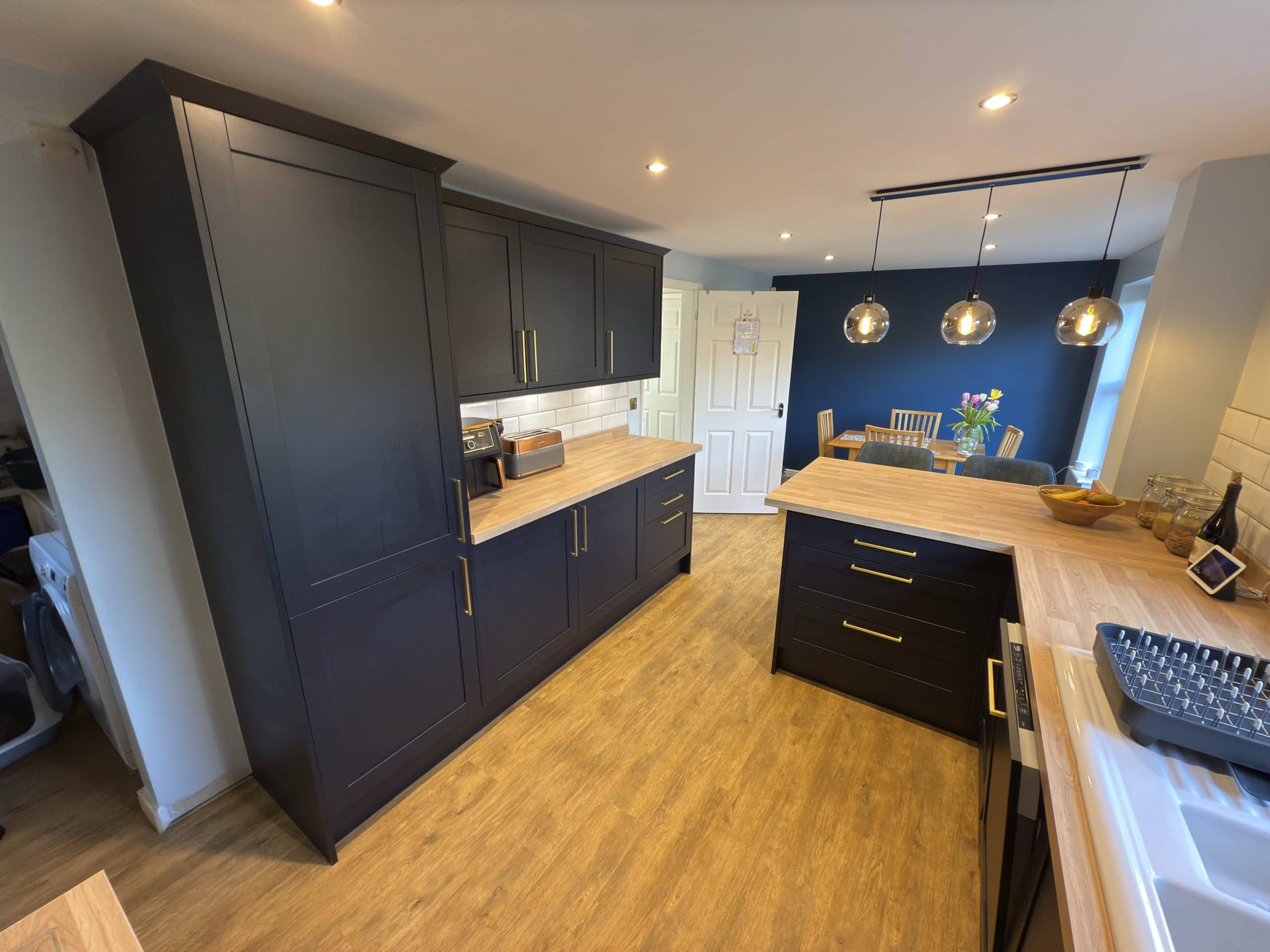 Modern kitchen and bathroom showroom design featuring dark cupboards, wooden worktops, and pendant lights above an island. In the background, a dining area with a table and flowers completes this stylish installation.