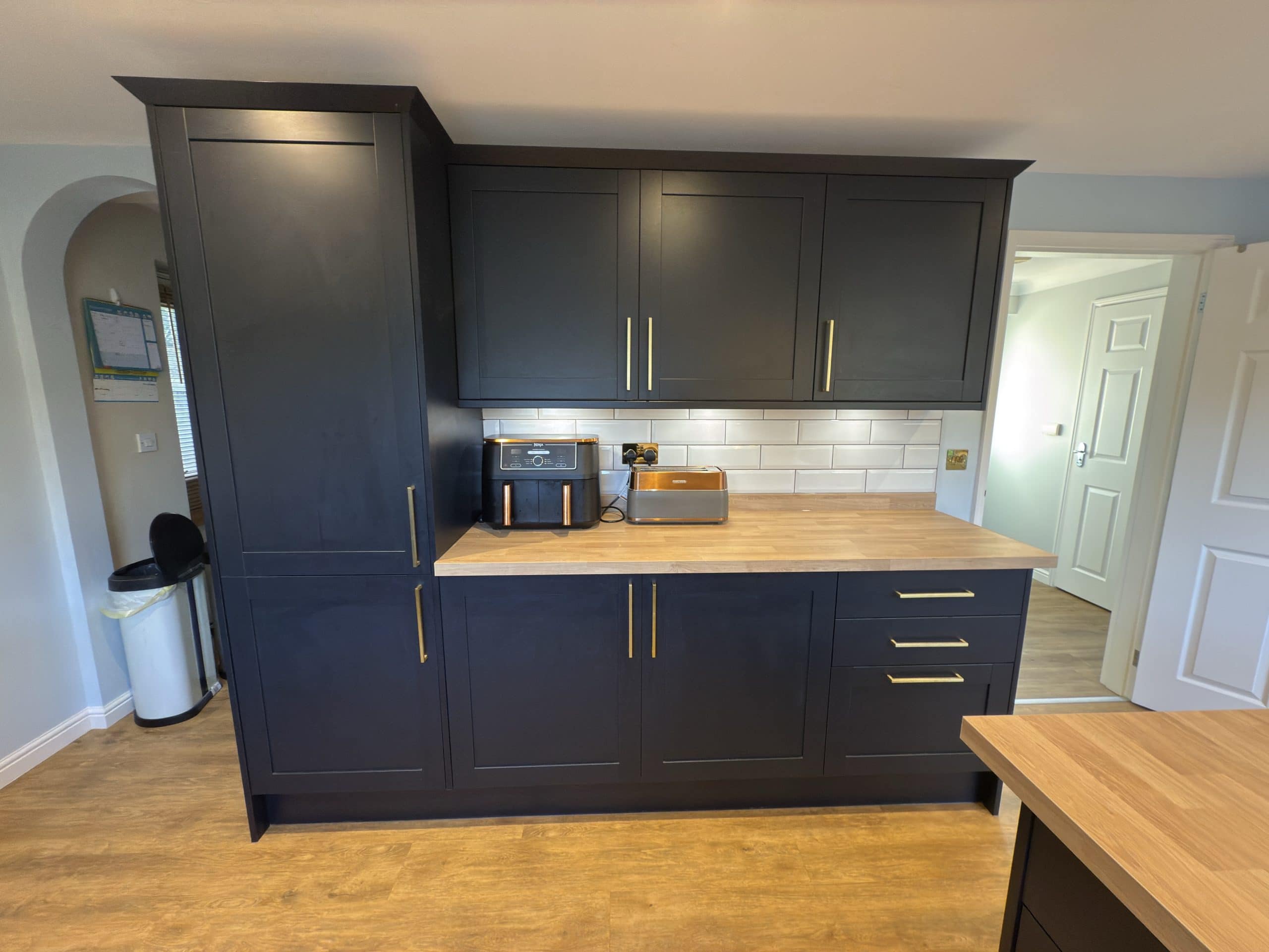 A modern kitchen with matt black cupboards, gold handles, wooden worktops, and a white subway tile splashback showcases expert showroom design. A toaster and air fryer sit on the counter; a white bin is in the corner beside the door.