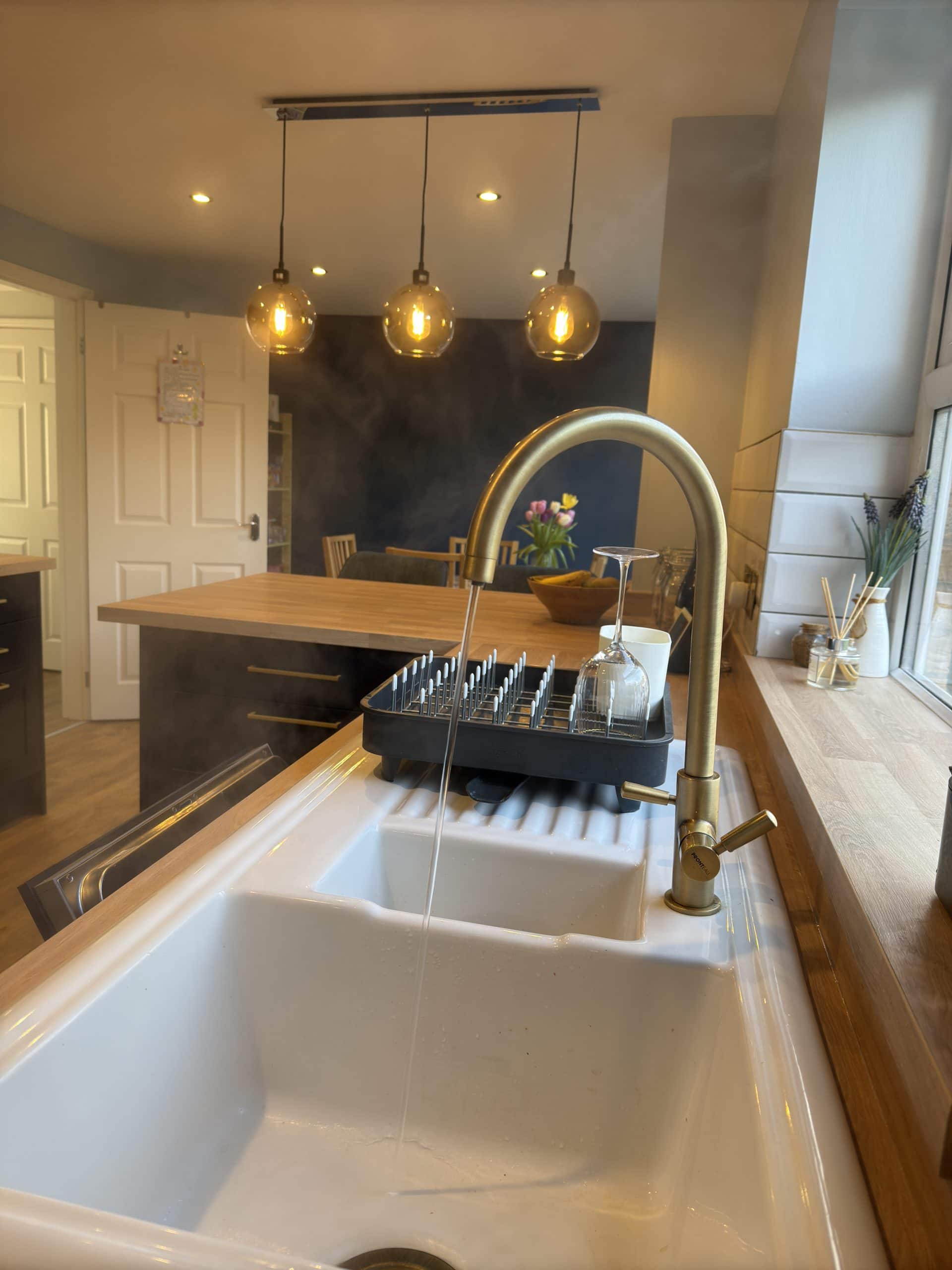 A modern kitchen designed with showroom-quality installation: white sink, brass tap running water, wooden worktop, and pendant lights. Steam rises near the tap; in the background, a dining table, chairs, and fruit bowl by the window.