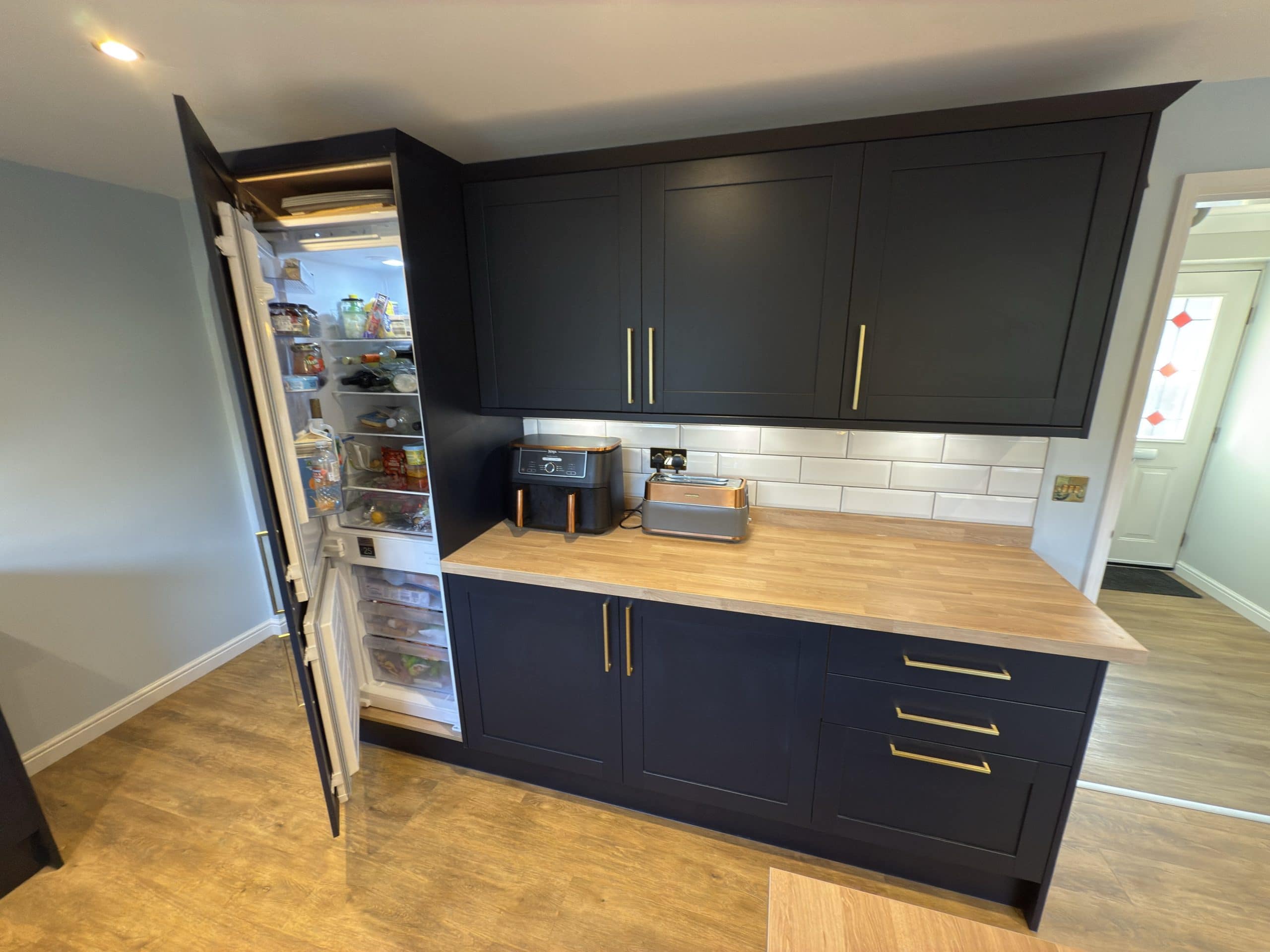 A modern kitchen with dark blue cupboards and brass handles, featuring a light wood worktop and white metro tile splashback. Perfect for kitchen and bathroom showroom inspiration or installation, with appliances neatly arranged on the counter.