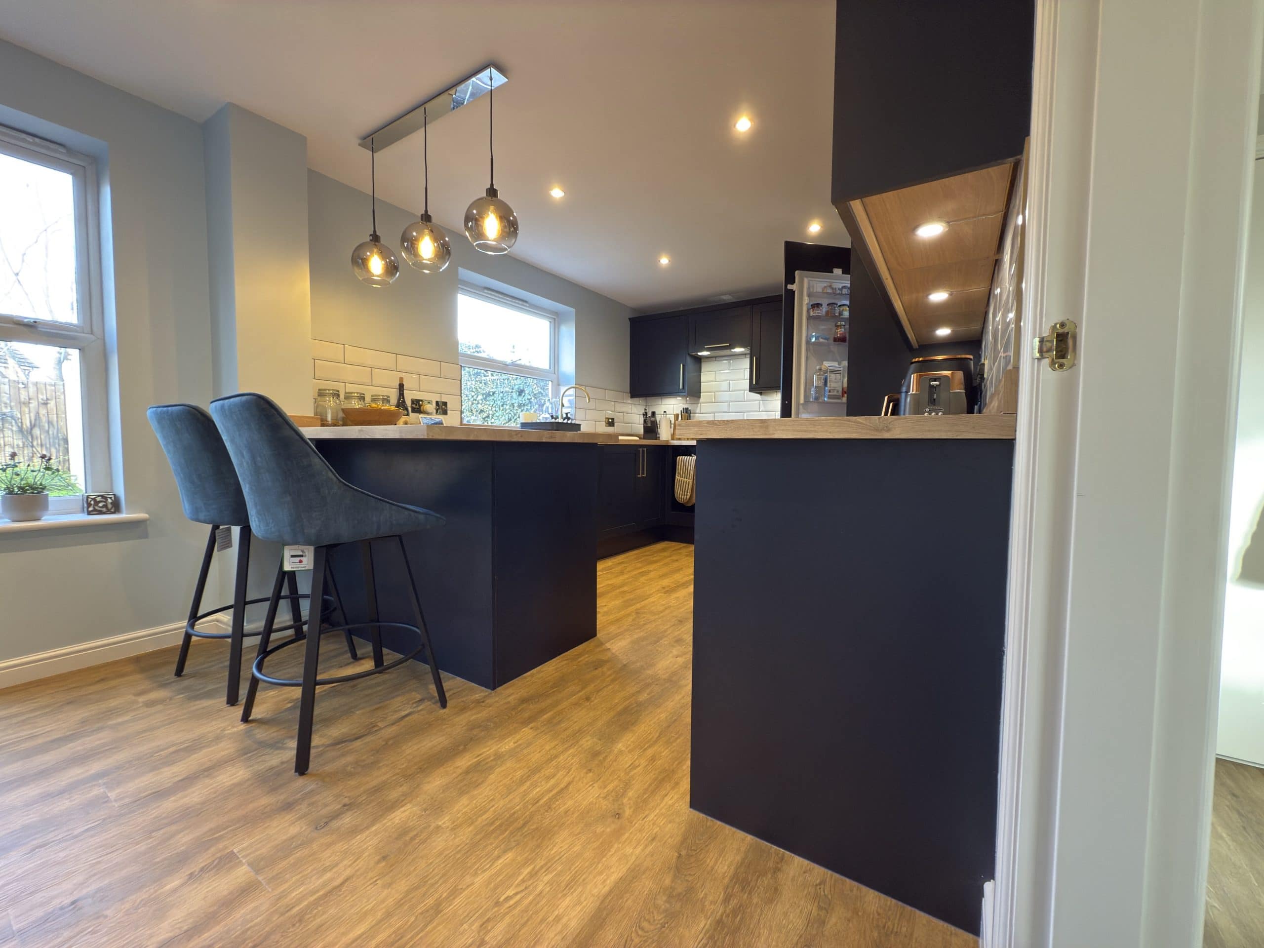 Modern kitchen and bathroom showroom-inspired design with black cupboards, wooden worktops, blue velvet bar stools, pendant lights, wooden flooring, and large windows. Fridge door is open, revealing contents inside.