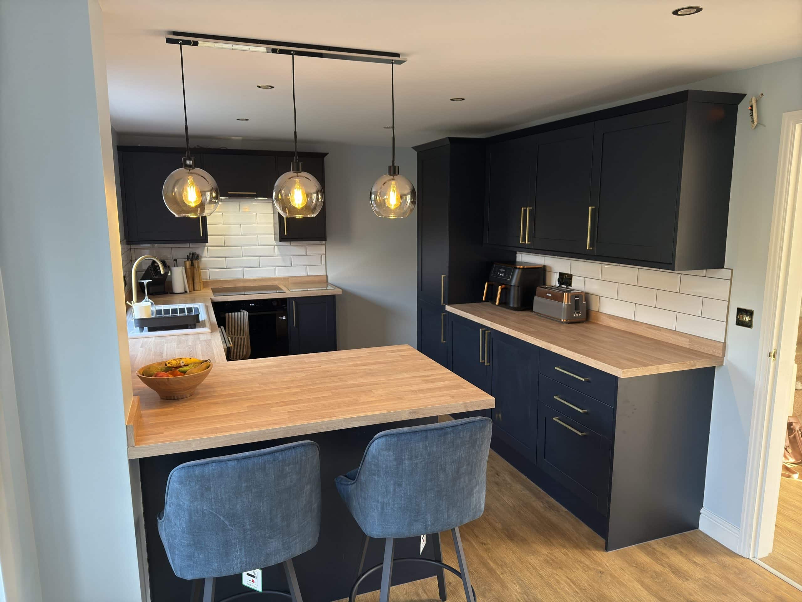 Modern kitchen and bathroom showroom design featuring navy blue cabinets, wooden worktops, white subway tile splashback, and stainless steel appliances. Pendant lights illuminate a breakfast bar with grey stools and a fruit bowl for an inviting touch.