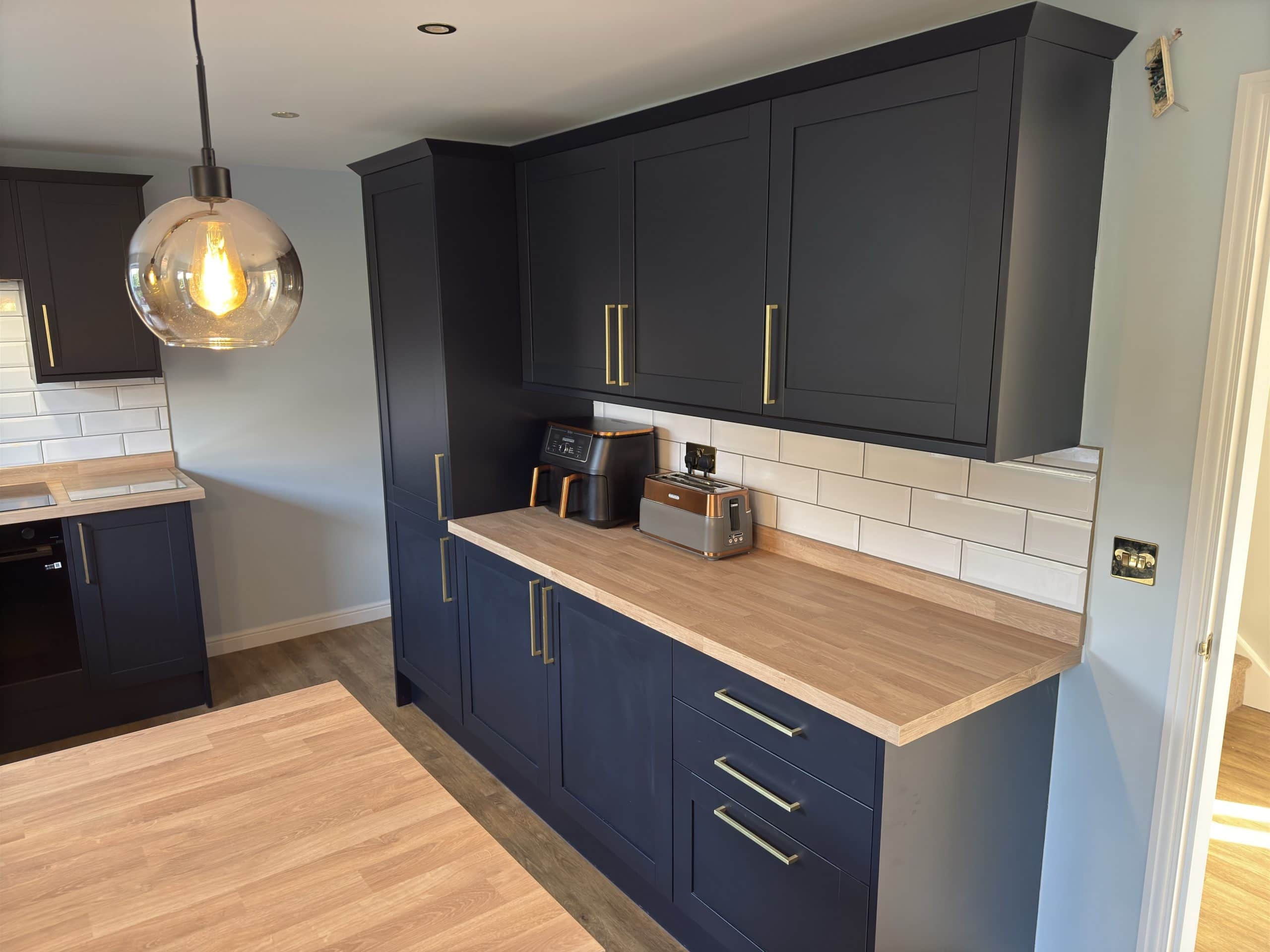 Modern kitchen and bathroom showroom-inspired design with navy blue cupboards, light wood worktops, white subway tile splashback, a toaster, air fryer, and a large glass pendant light hanging from the ceiling.