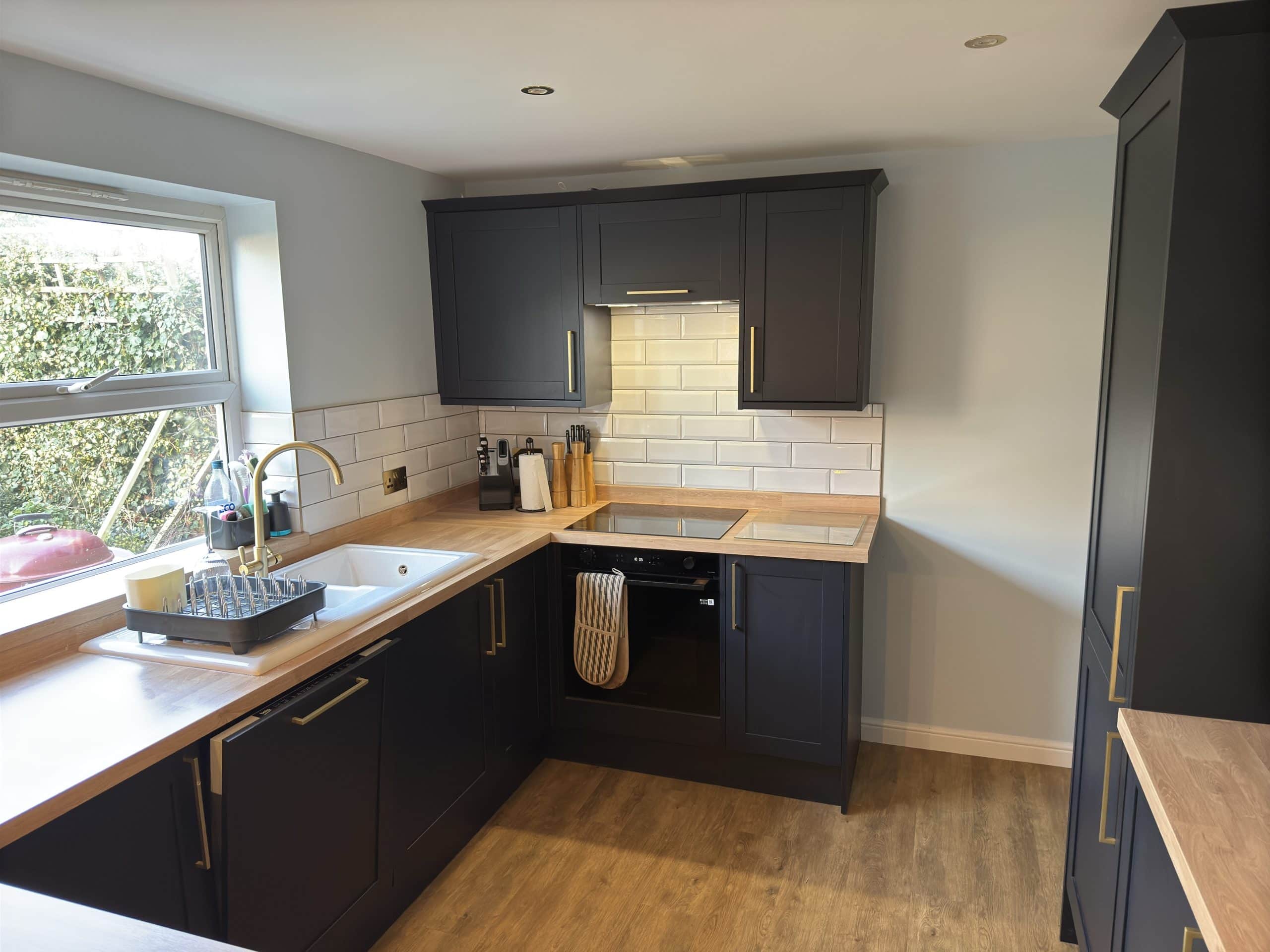 Modern kitchen with dark cupboards, light wooden worktops, and a white metro tile splashback. Ideal for kitchen and bathroom showroom installation, its design features an electric hob, sink by the window, and natural light brightening the space.