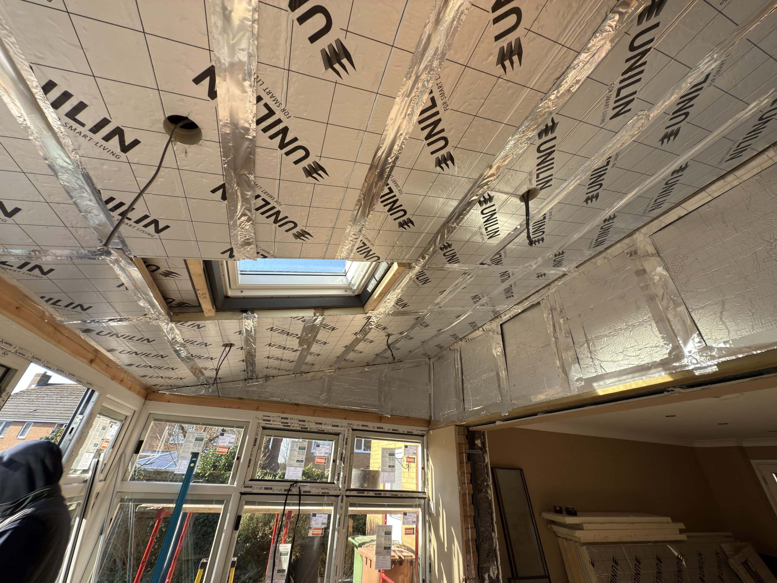 The image shows a partially renovated room with foil-backed insulation panels on the ceiling, ideal for kitchen and bathroom showroom installation. A skylight centres the space, sunlight streams in, and windows line one wall amid construction materials.