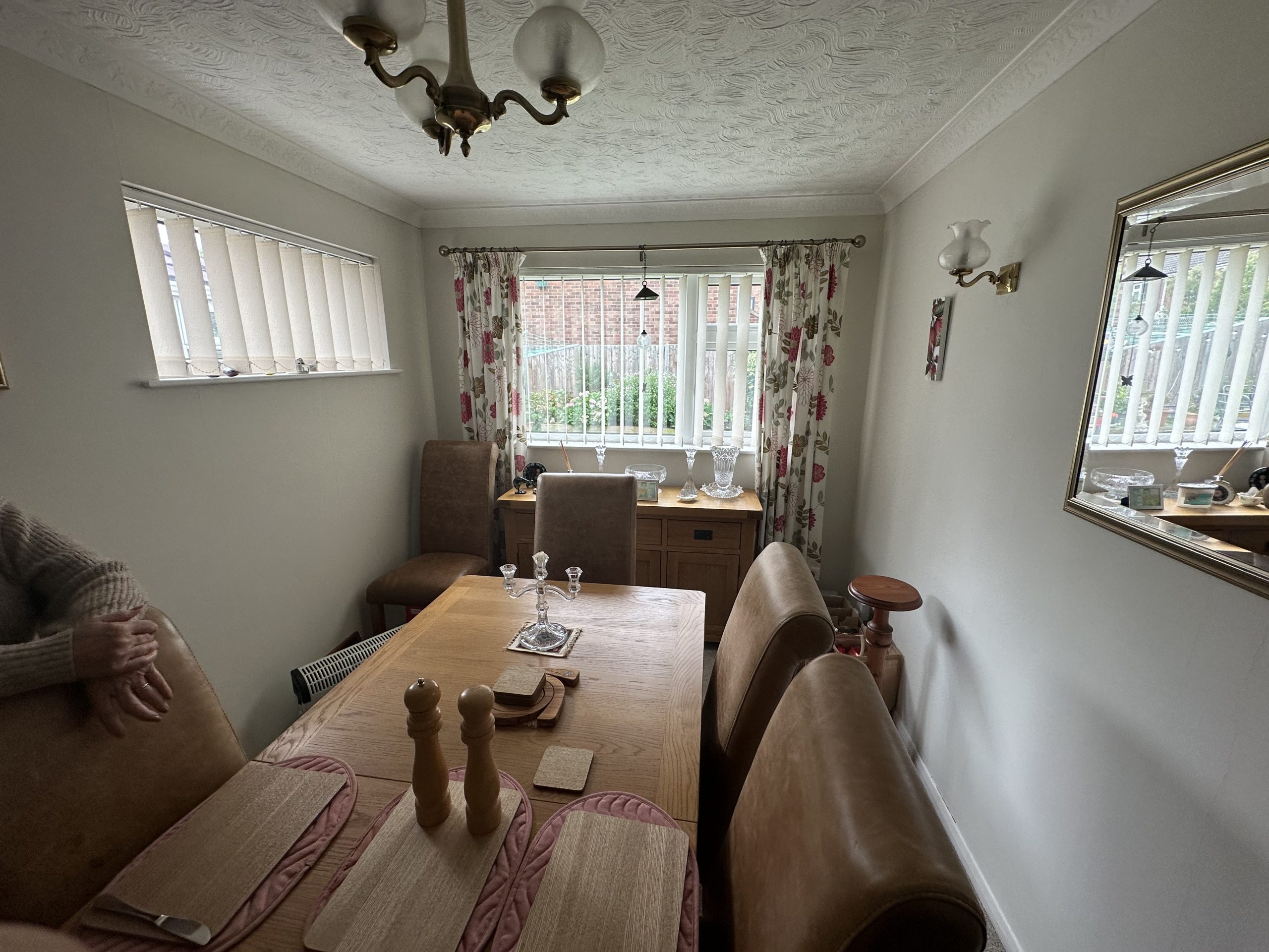 A dining room with a wooden table set for four, brown leather chairs, placemats, and pepper mills offers a warm space. Large windows with blinds and floral curtains echo thoughtful design found in a kitchen and bathroom showroom. A person's arm rests at the table's edge.