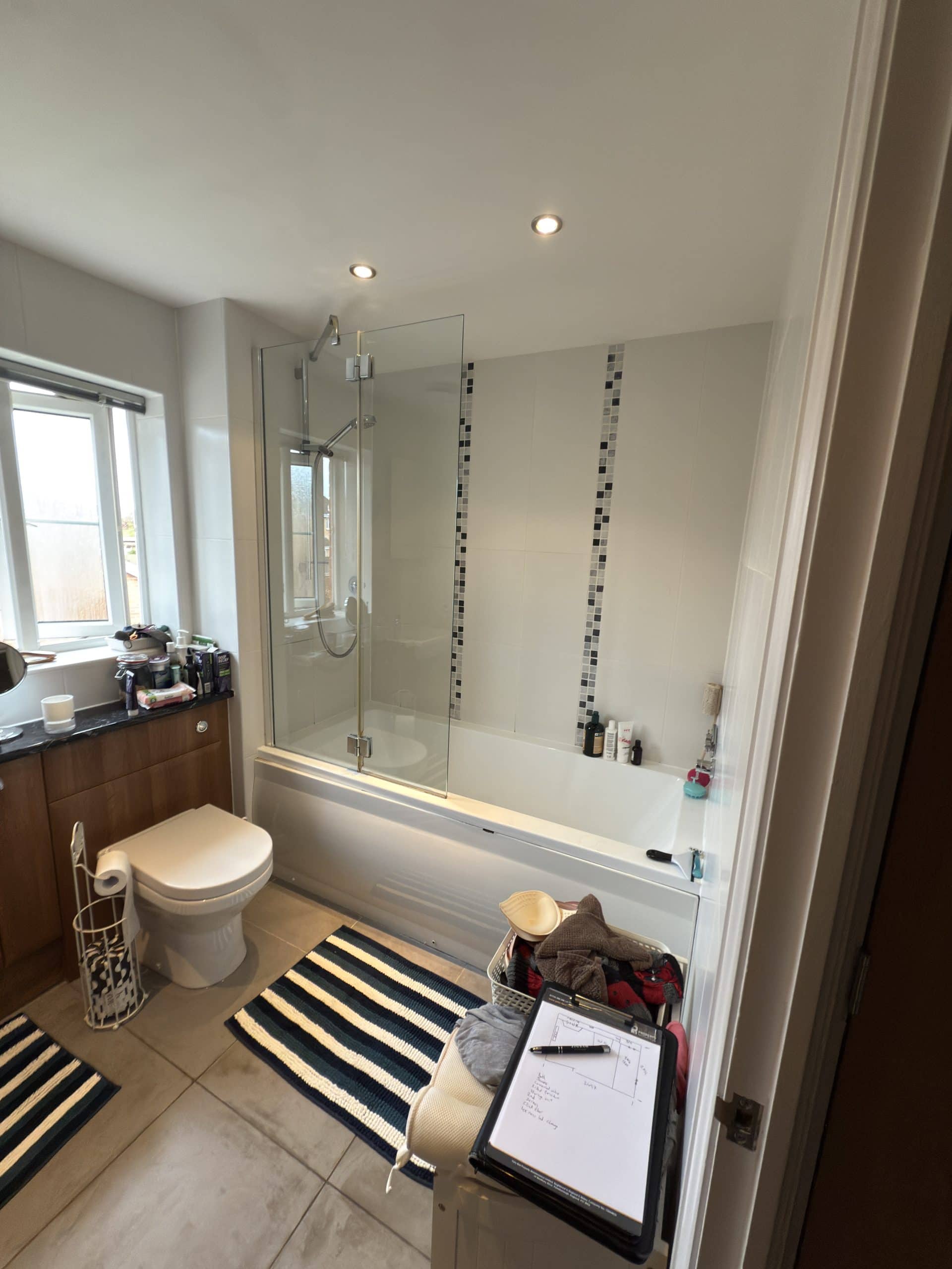 A modern bathroom designed with a sleek bath, glass shower screen, stylish striped bath mats, and quality fittings—perfect inspiration from a kitchen and bathroom showroom. A laundry basket and whiteboard sit conveniently near the door.