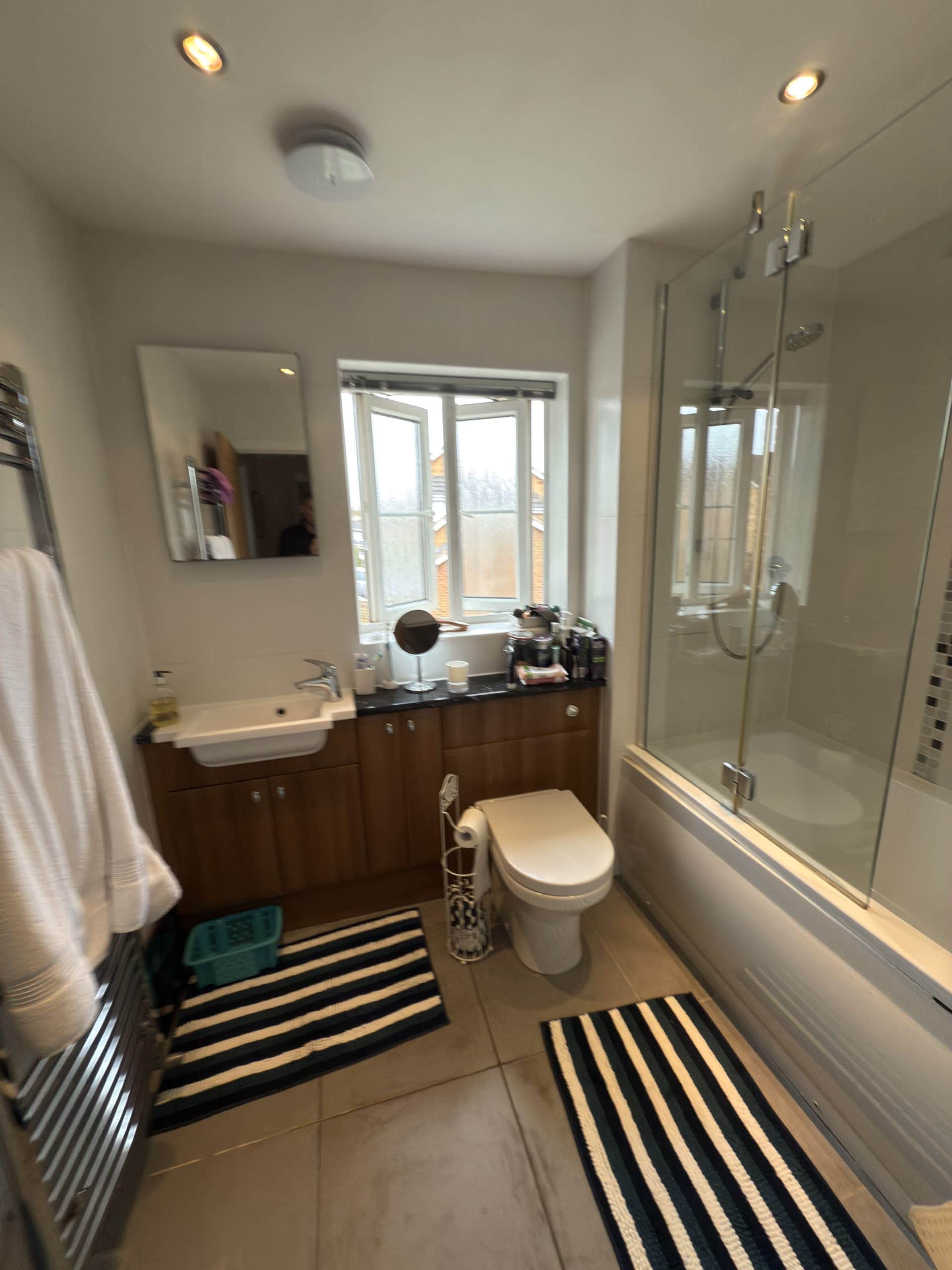 A modern bathroom designed with a white basin, toilet, and bath with a glass screen. Wooden cupboards from a kitchen and bathroom showroom brighten the space, while striped rugs and an open window add style. Towels hang on a rail by the door.
