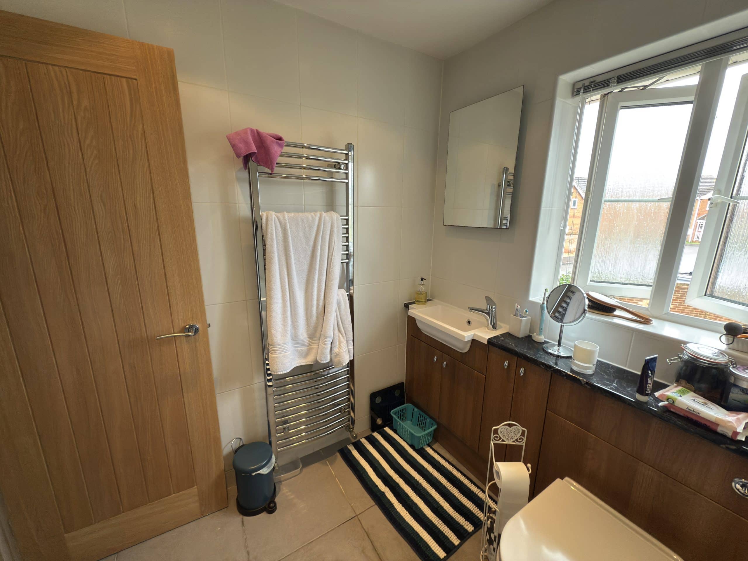 A bathroom with a wooden door, heated towel rail with white towels, striped rug, washbasin with cupboard, mirror, window, and various toiletries on a dark worktop—showcasing expert bathroom showroom design and installation.
